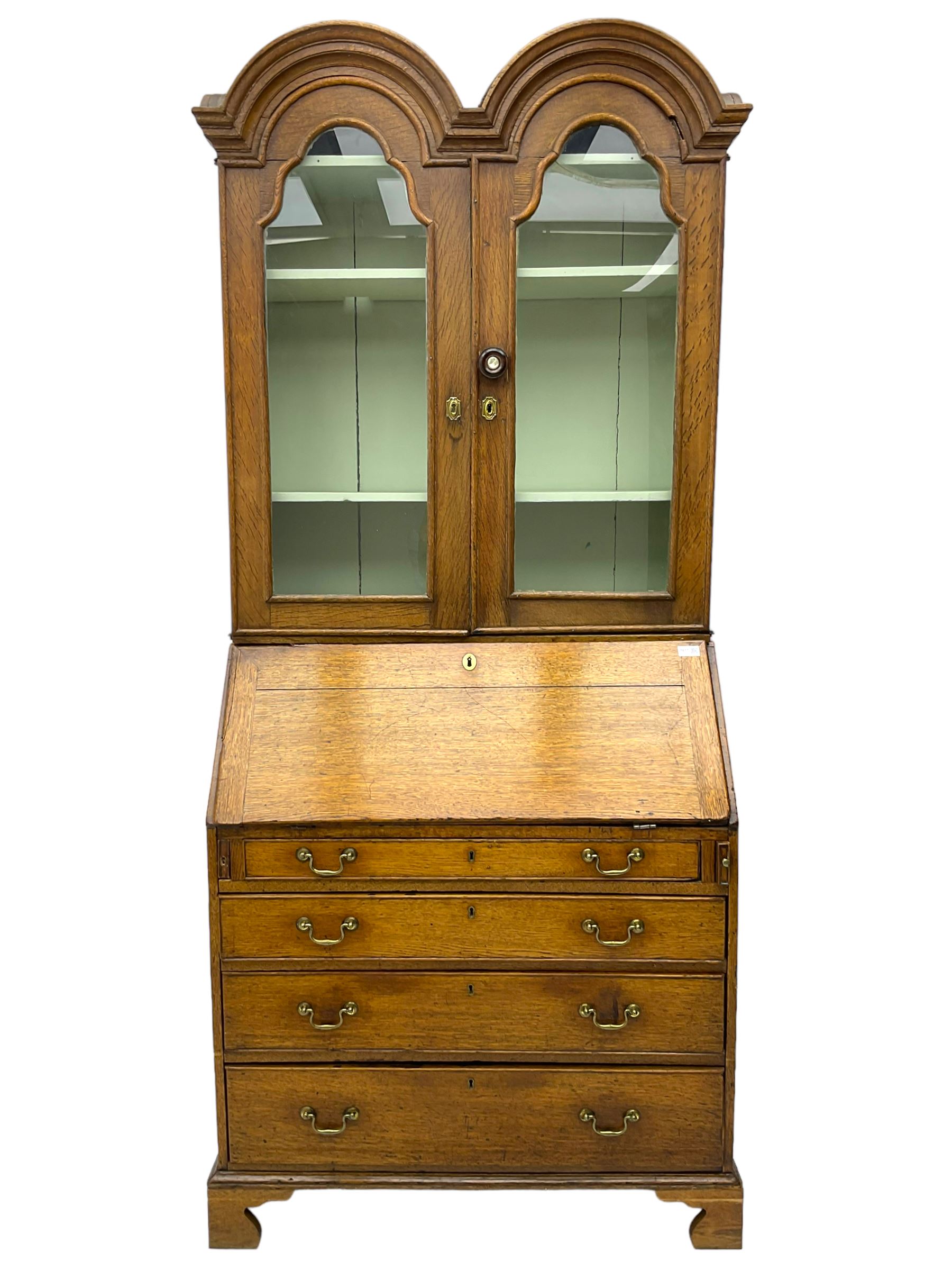 George III oak bookcase on bureau, the double arched top enclosed by two shaped glazed doors, fall-front concealing fitted interior, over four graduating cock-beaded drawers, on bracket feet
