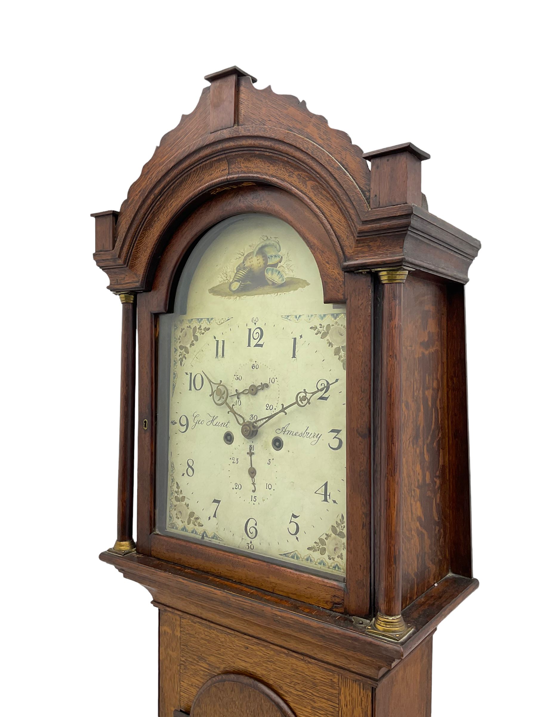 A provincial oak longcase clock retailed by “Geo Hunt, Amesbury” c1820, with a crested break arch pediment and corresponding glazed hood door flanked by two plain pillars with capitals, long trunk with a full length break arch door on a rectangular plinth with a shaped base, painted dial with Arabic numerals, minute markers, subsidiary date and seconds dials and stamped brass hands, matching painted spandrels and a depiction of sea shells to the arch, dial pinned to the movement via a Walker Hughes cast falseplate, with an eight day rack striking movement, striking the hours on a cast bell. With weights and Pendulum




