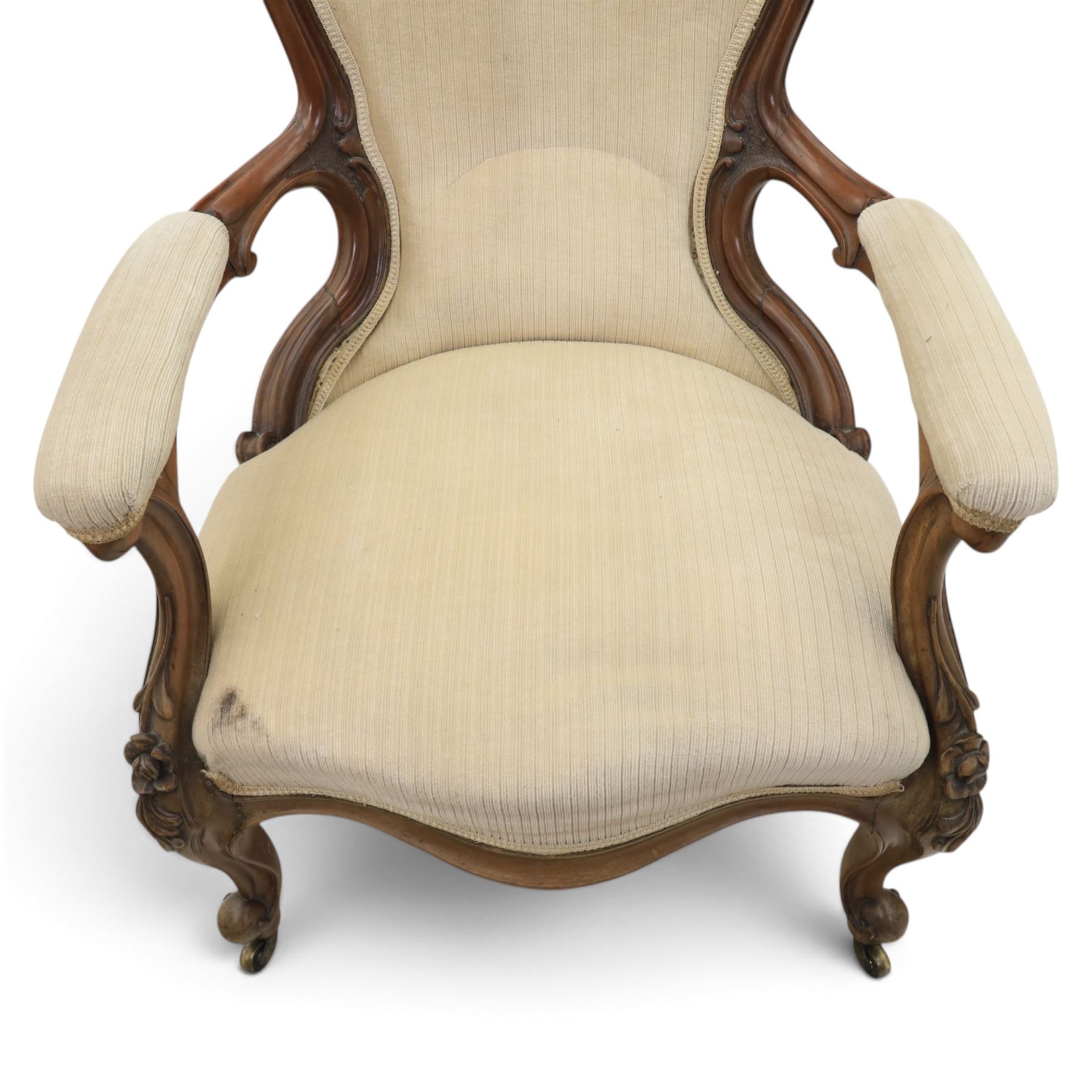 Late Victorian walnut open armchair, the carved shaped cresting rail centred by scrolls and foliage above a padded serpentine back and overstuffed seat upholstered in cream striped fabric, with padded scroll arms on open carved supports, raised on cabriole supports with carved knees and ceramic castors