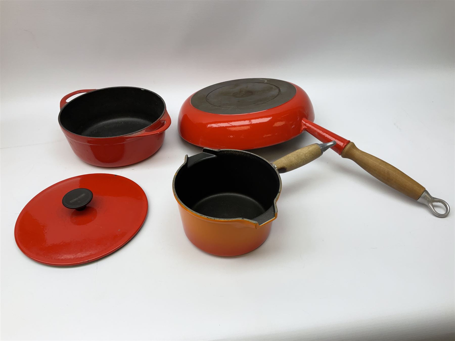 Le Creuset 'Volcanic Orange' cast iron and enamel milk pan with wood handle together with a three piece red cast iron Cousances saucepan set of graduating form, with lids housed in original wooden hanging stand and further Cousances lidded casserole dish and pan