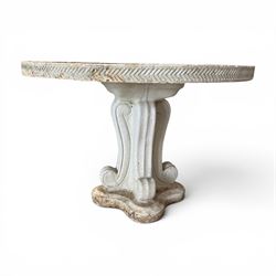 Weathered cast stone garden table, circular top etched with central floral medallion, the edge with repeating chevron motifs, raised on a quadriform S-scroll pedestal with stepped and chevron carved base