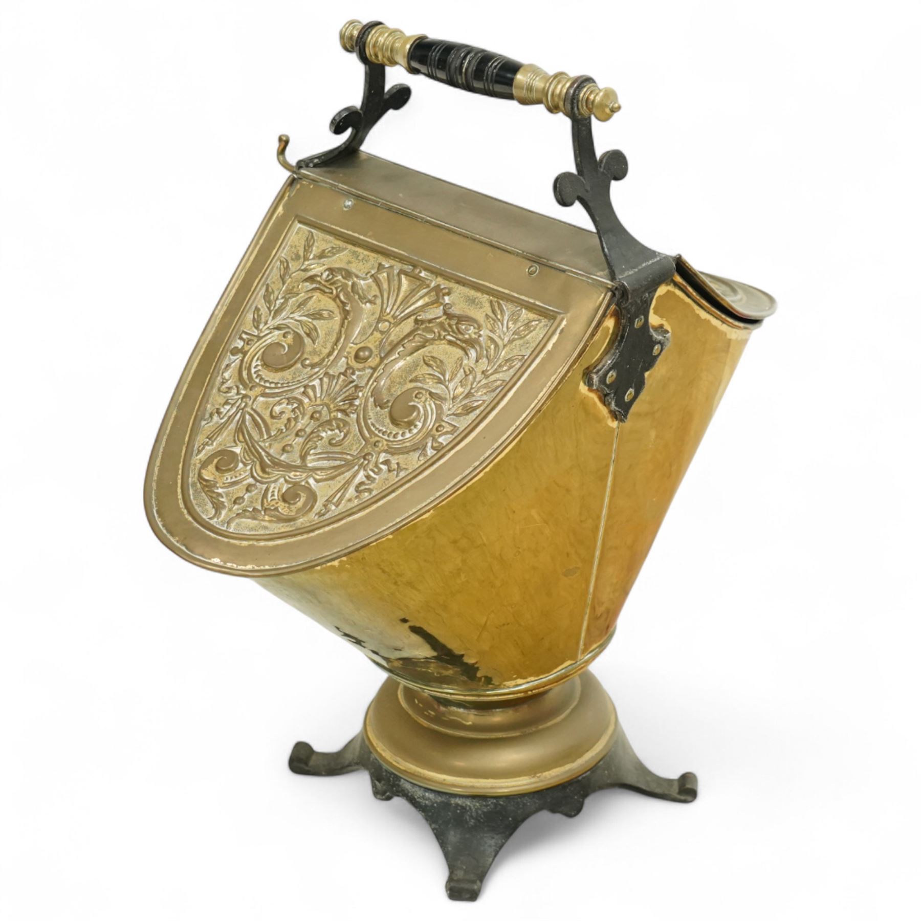 Late 19th century brass coal scuttle, of helmet form, with embossed scrolling foliate panels, cast iron supports with turned ebonised handle, raised on a cast iron tripod base