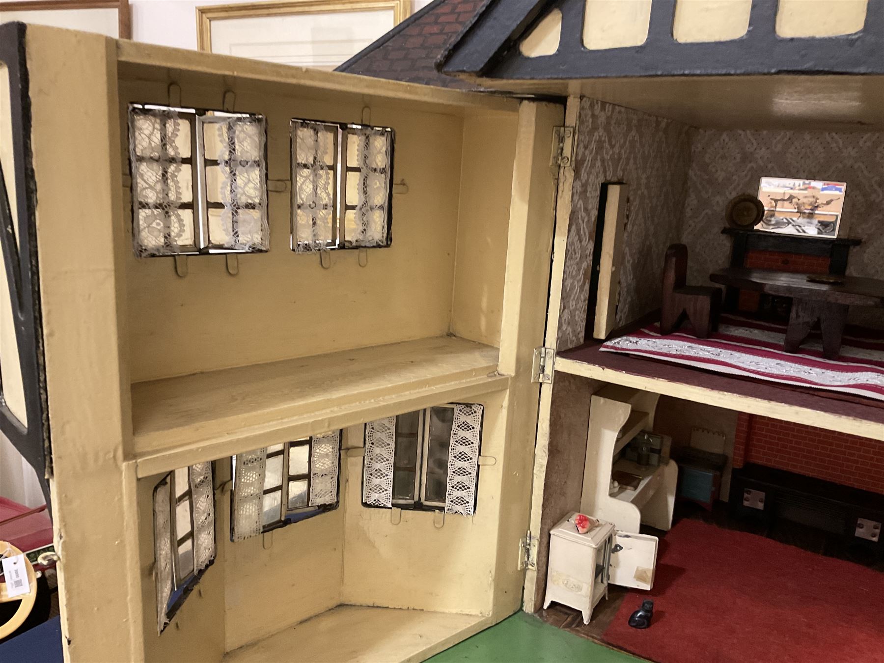 Large Tri-ang No.93 doll`s house in the 'Stockbroker's Range' c1935; two-storey double-fronted Tudor design, half timbered gables, two-storey bay windows, cream and mock shrubbery to front, opening metal framed windows with green shutters, red simulated tiled roof with chimney, integral garage with opening doors, front elevation has four hinged doors opening to reveal two bedrooms with fire places, living room, bathroom and kitchen with built in dresser and sink, entrance hall and staircase, side entrance with porch and seat and sun dial over on chimney stack; some wiring for electrical lighting; metal Tri-ang Toys disc verso; comprehensively furnished H66cm L117cm D43cm