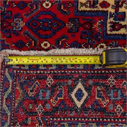 North West Persian Senneh hand-knotted rug, dark indigo field with a central bold stepped lozenge medallion of madder red and deep blue, enclosed within an ivory serrated contour forming a cruciform outline, medallion with repeating boteh and floral devices, field decorated with an all-over Herati pattern of rosettes enclosed by angular leaves principal border of madder red ground decorated with alternating floral heads, flanked by narrow guard stripes, with cotton fringe ends 