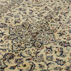Central Persian Kashan hand-knotted rug, ivory ground, with a small central floral medallion and pendants, the field finely decorated with blue, cream and soft red scrolling vines, flowerheads and palmettes, within an ivory floral border and narrow guard stripes, with cotton fringes