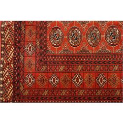 Turkmen hand-knotted carpet, madder red field, with an all-over design of row of Tekke guls, each enclosing stylized cruciform and geometric devices in tones of ivory, dark brown, and indigo, principal border of stylized geometric motifs, flanked by guard border with angular latch hook geometric designs, with cotton fringe ends 
