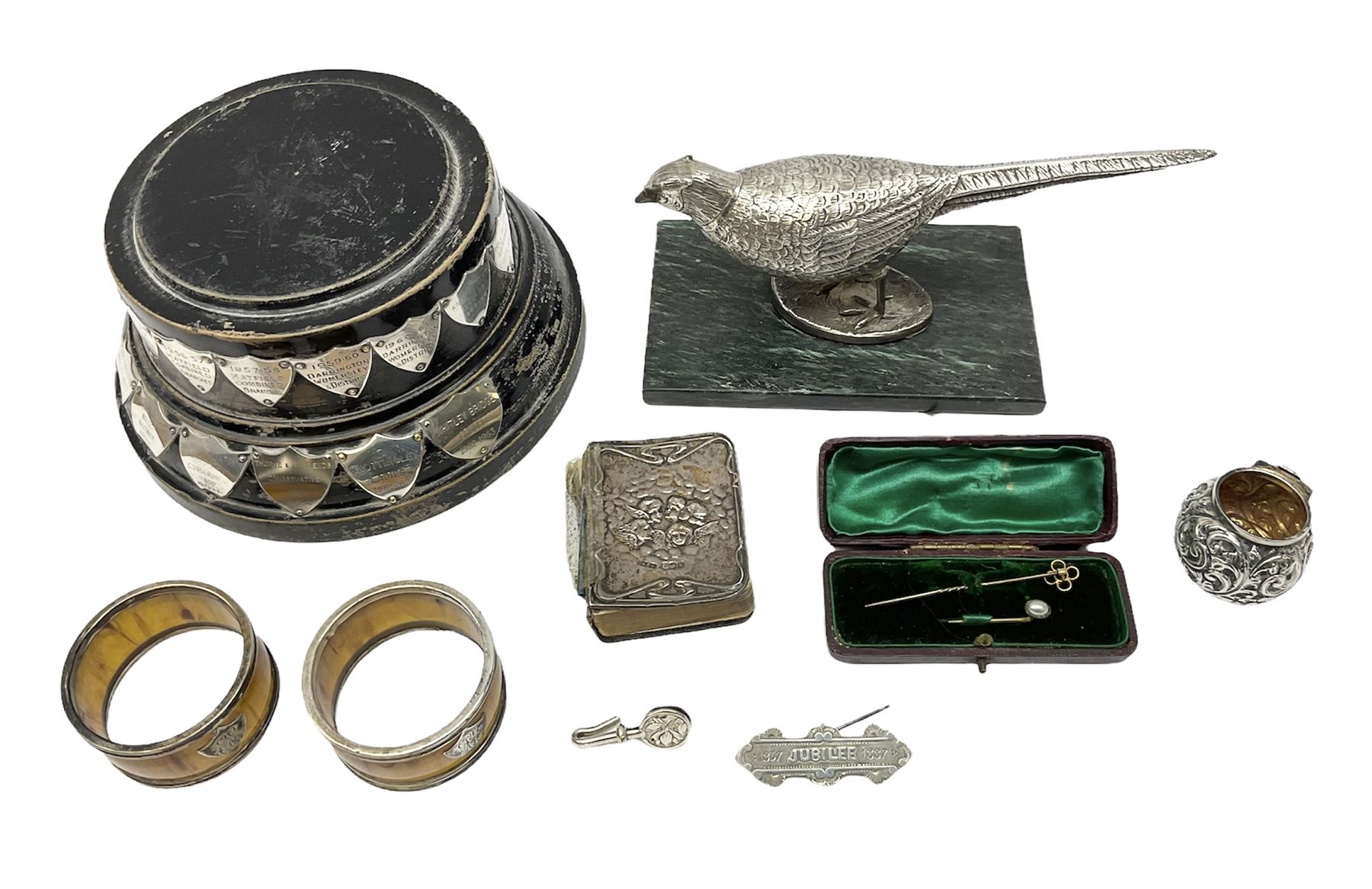 Group of silver, to include filled figure of a pheasant, hallmarked for London, pair of silver mounted horn napkin rings, miniature Edwardian bible with silver cover embossed with putti, Victorian silver brooch, ebonised plinth with applied inscribed silver shields, etc., together with two stick pins, one example with 9ct gold terminal 