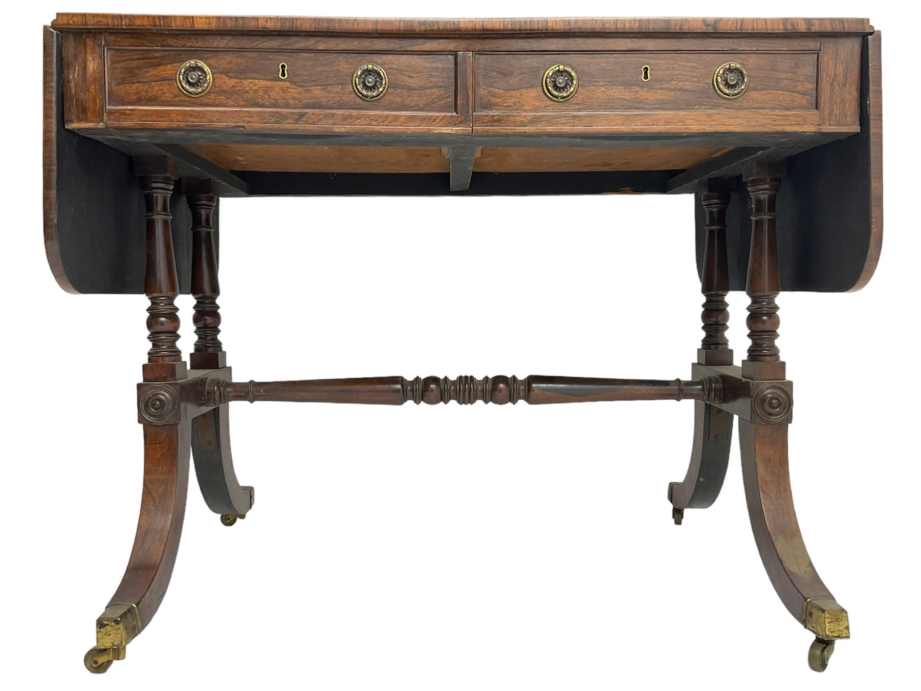 Regency rosewood sofa table, drop leaf rectangular top with rounded corners, fitted with two drawers and two opposing false drawers, raised on dual turned columns terminating in splayed feet united by turned stretcher, on brass cups and castors