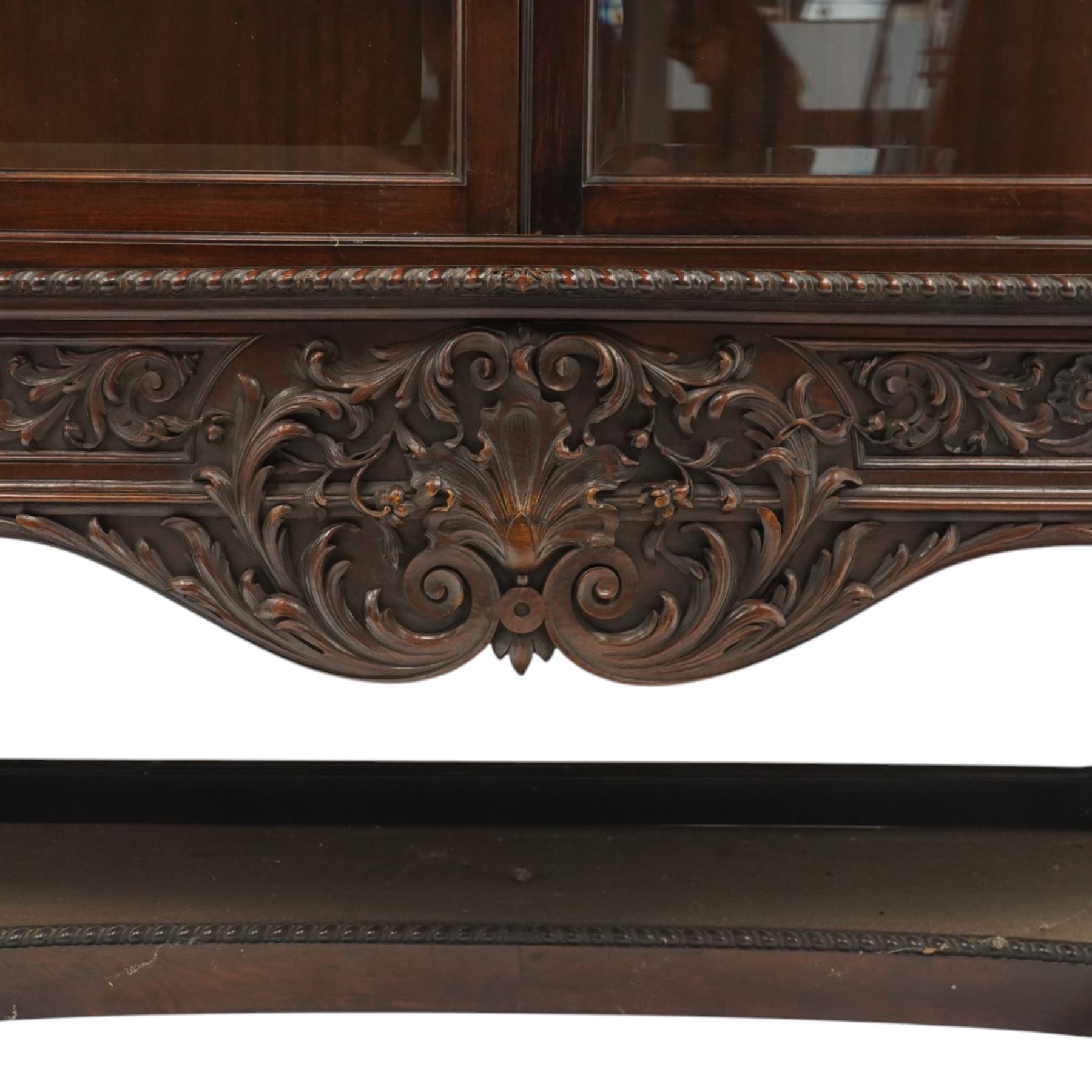 Large and impressive 19th century heavily carved mahogany display cabinet, the ornate pediment carved with a central scalloped and foliate rocaille motif with extending acanthus leaves and egg-and-dart moulding flanked by acanthi finials, over a central cartouche with carved bow-and-arrow motif and trailing scrollwork, the bookcase enclosed by two glazed doors with bevelled glass sides, the base with a gadrooned upper edge over a central rocaille ornament with scrolling acanthus leaves, the panelled frieze with further foliate motifs, raised on large carved snarling lion mask supports with floral garlands and claw feet, united by a concave rectangular platform base with gadrooning egg-and-dart moulding