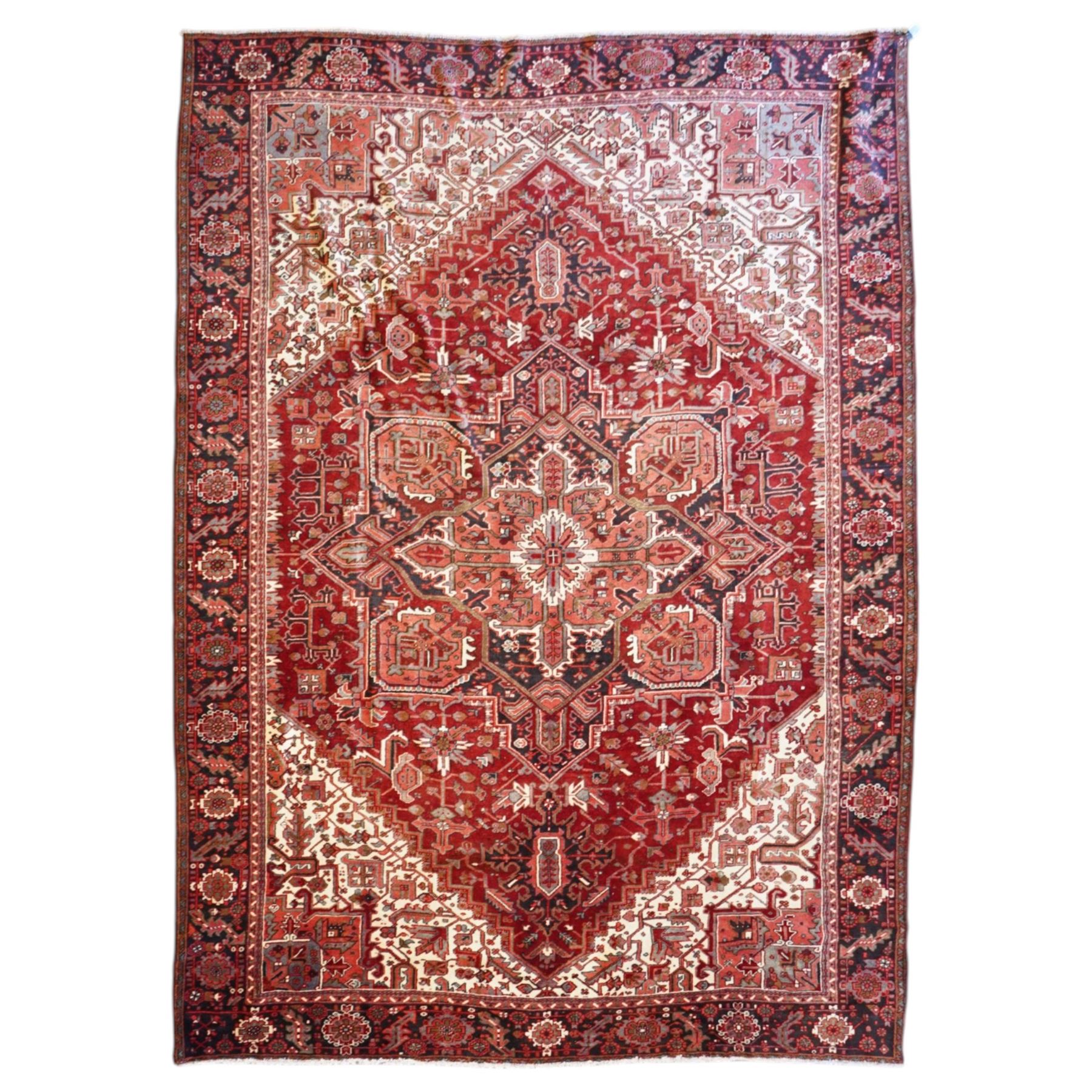 North West Persian Heriz hand-knotted carpet, brick red field with a large central angular star medallion with pendants surrounded by stylized geometric palmettes, serrated leaves and tribal motifs,, ivory corner spandrels, field enclosed by a dark indigo main border of stylize rosettes and angular vine motifs between multiple guard stripes, with cotton fringe ends 