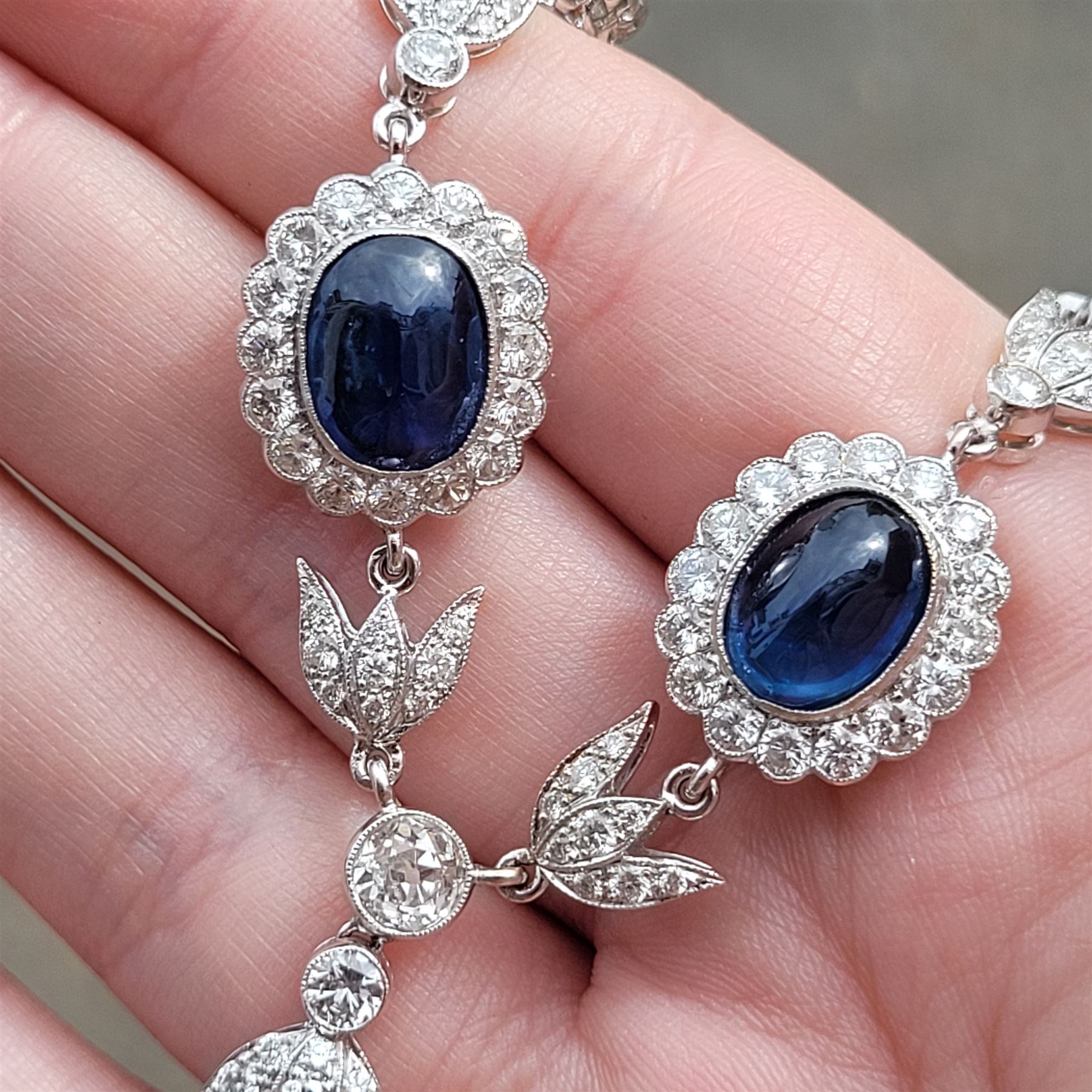 18ct white gold sapphire and diamond milgrain set necklace, oval cabochon cut sapphire surrounded by eight round brilliant cut diamonds and eight French cut sapphires, suspending from a diamond set leaf pendant, single round brilliant cut diamond and a larger old cut diamond, the chain set with six graduating cabochon cut sapphires, each surrounded by diamonds and spaced by a single diamond and a diamond set leaf, to a belcher link chain necklace, hallmarked, total sapphire weight approx 27.00 carat, total diamond weight approx 7.50 carat