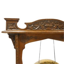 Late Victorian Arts & Crafts carved oak framed floor standing dinner gong, the cresting rail carved with central urn and trailing foliage, over shaped end supports decorated with graduating tulip and floral motifs, on rectangular base with scroll sledge feet, the brass gong stamped with William Suckling of Birmingham 'Burmese' makers mark, with striker