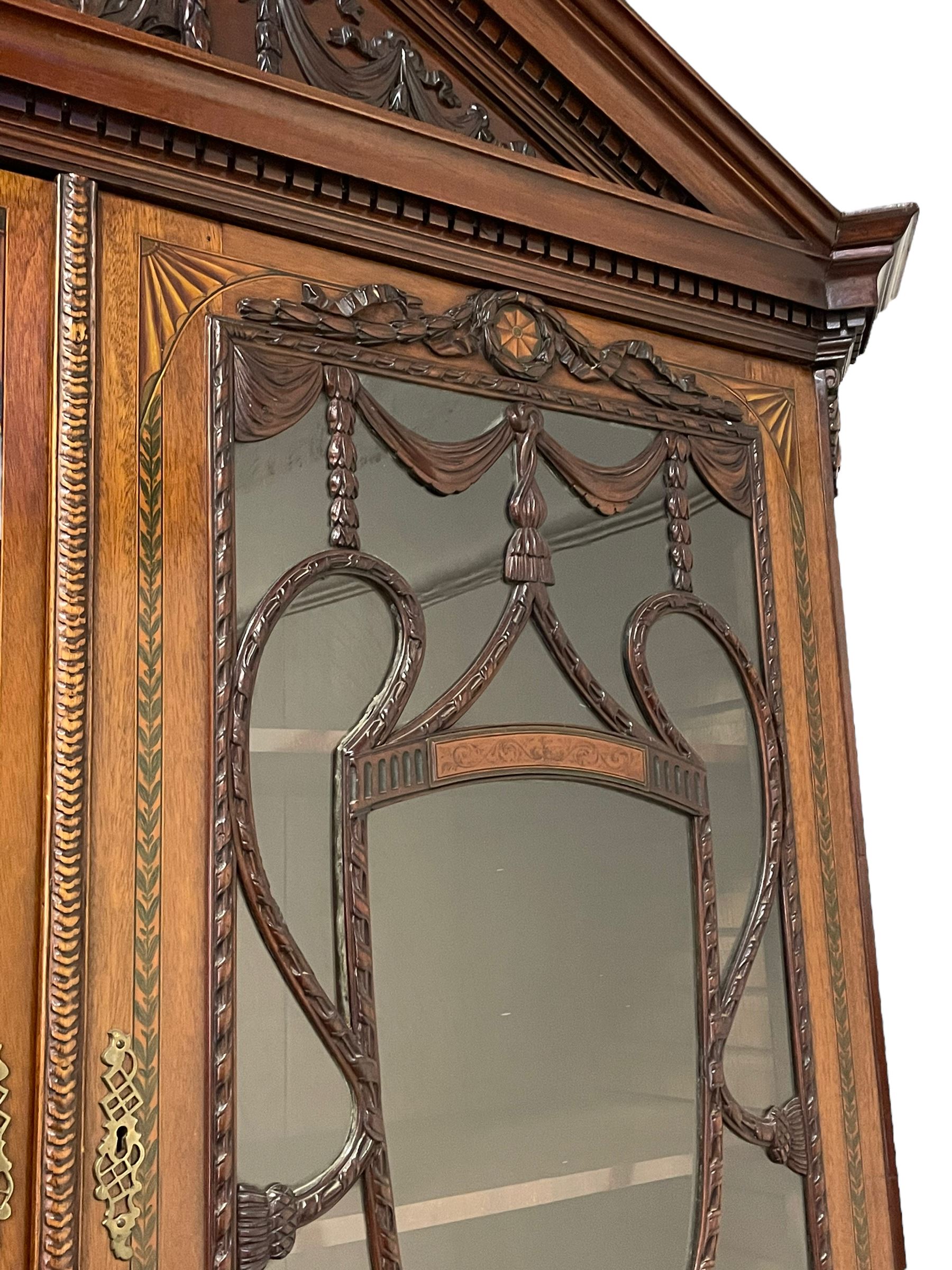 Large and impressive late 19th century Irish mahogany secretaire bookcase, moulded sloped arch dentil pediment decorated with central carved mask surrounded by linen festoons and trailing bellflowers, canted and fluted uprights with carved acanthus leaf upper and lower brackets, enclosed by two astragal glazed doors in the form of trophies with twist ribbon carved mouldings, inlaid with fanned spandrels and trailing leaf painted satinwood band, cylinder roll top inlaid with central fan motif and fanned spandrels, pull-out action revealing stepped fitted interior, central tambour roll cupboard flanked by two Composite pilasters and a combination of drawers and pigeon holes, fitted with three break-front drawers, matched canted and fluted corners with acanthus leaf carvings, on square tapering and fluted spade feet