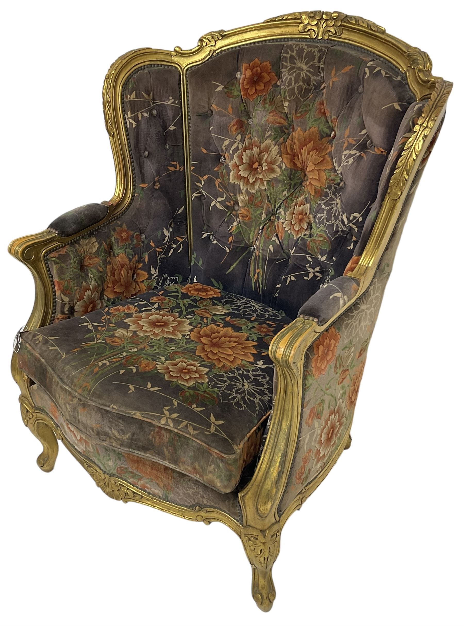 Pair of Louis XVI design gilt framed wingback armchairs, cresting rail carved and moulded with foliate decoration and C-scrolls, upholstered in buttoned floral patterned mauve velvet with loose seat cushion, scallop carved apron over cabriole supports