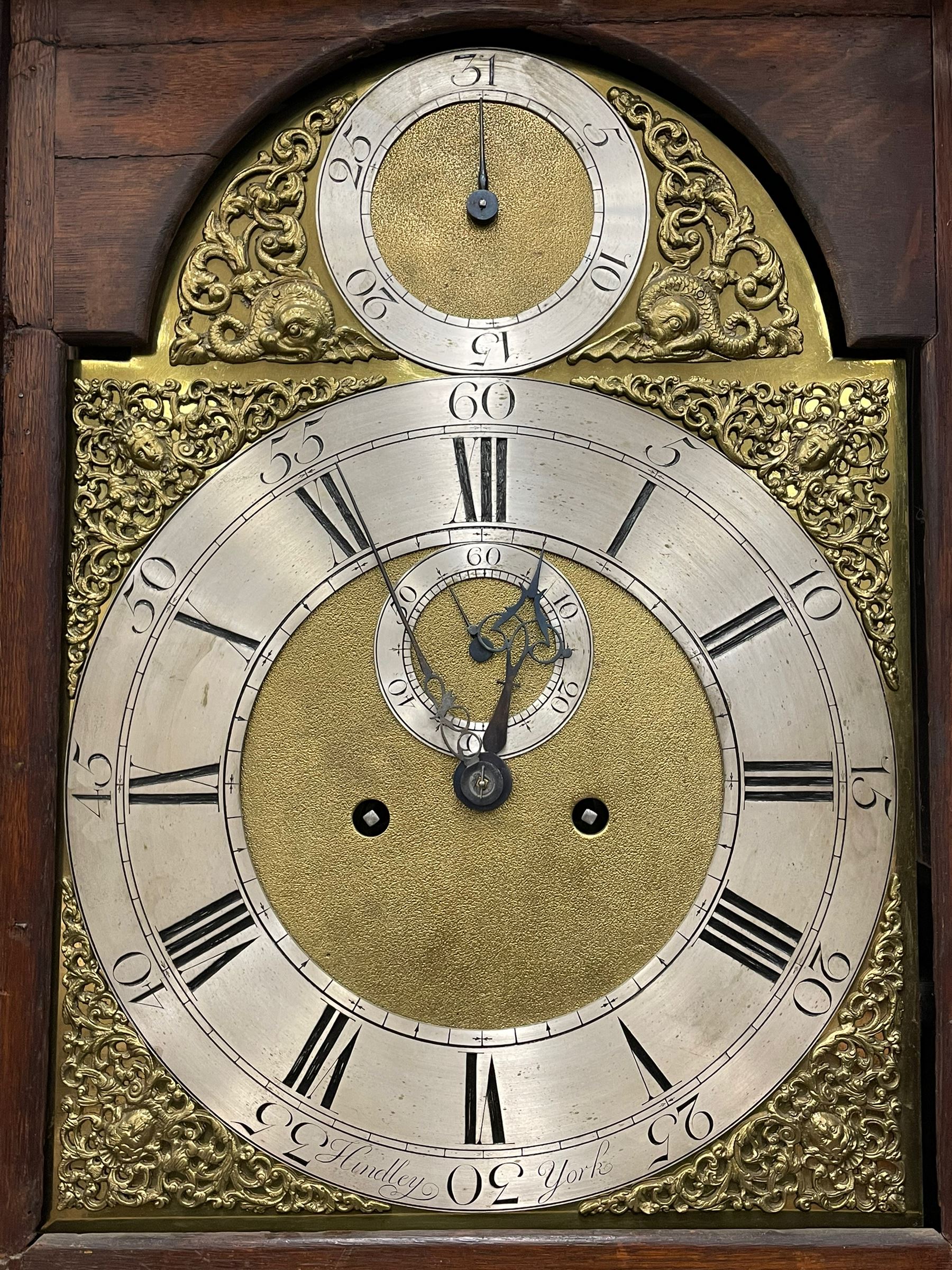 Henry Hindley of York – mid-18th century oak cased 8-day longcase clock,
flat topped caddy pediment with gesso finials, rectangular hood door flanked by wooden turned pilasters with capitals, canted trunk with a full-length break arch topped door on a short plinth with applied skirting and bracket feet, brass dial with a silvered chapter ring, Roman numerals and five-minute Arabic’s, minute and inner quarter hour tracks, matted dial centre with subsidiary seconds dial, blued steel hands and female head pattern spandrels, silvered calendar dial to the break arch with dolphin spandrels, four pillar rack striking movement, striking the hours on a bell. With weights and pendulum. 
H250 W48 D27
Henry Hindley originally from Wigan (Lancs) is recorded as working in York from Petergate 1731-41 then Stonegate 1741-1771. A famous and highly innovative maker, often referred to as the “Tompion of the North.” 
