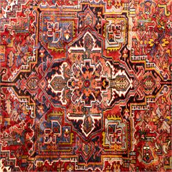 North West Persian Heriz hand-knotted carpet, madder red ground, with a large central geometric medallion with pendants, surrounded by angular foliate and floral motifs incorporating stylised rosettes, palmettes and serrated vine work, enclosed by ivory spandrels and a dark navy border with repeating geometric flowerheads and angular meandering vines, flanked by narrow patterned guard stripes, with cotton fringes
