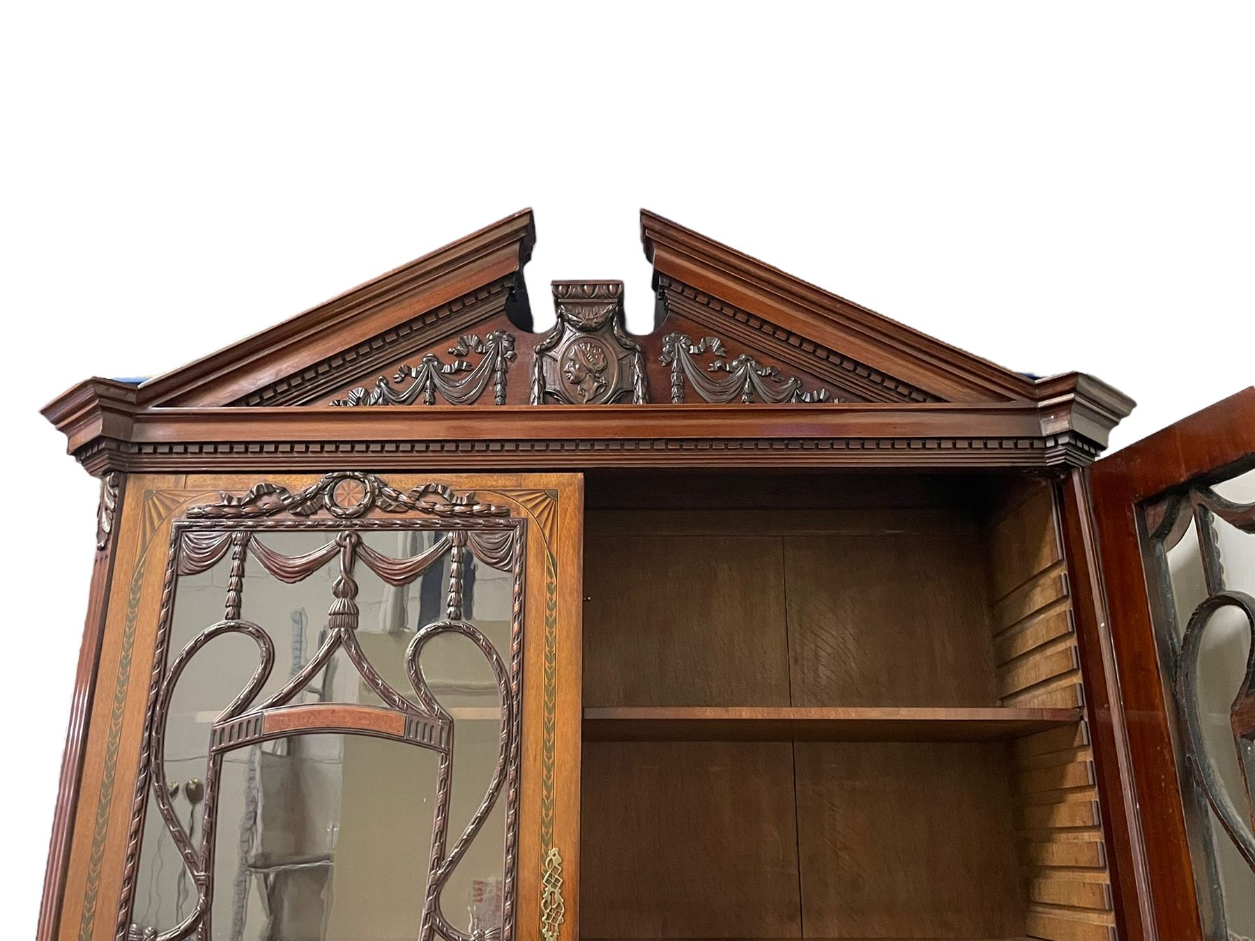 Large and impressive late 19th century Irish mahogany secretaire bookcase, moulded sloped arch dentil pediment decorated with central carved mask surrounded by linen festoons and trailing bellflowers, canted and fluted uprights with carved acanthus leaf upper and lower brackets, enclosed by two astragal glazed doors in the form of trophies with twist ribbon carved mouldings, inlaid with fanned spandrels and trailing leaf painted satinwood band, cylinder roll top inlaid with central fan motif and fanned spandrels, pull-out action revealing stepped fitted interior, central tambour roll cupboard flanked by two Composite pilasters and a combination of drawers and pigeon holes, fitted with three break-front drawers, matched canted and fluted corners with acanthus leaf carvings, on square tapering and fluted spade feet