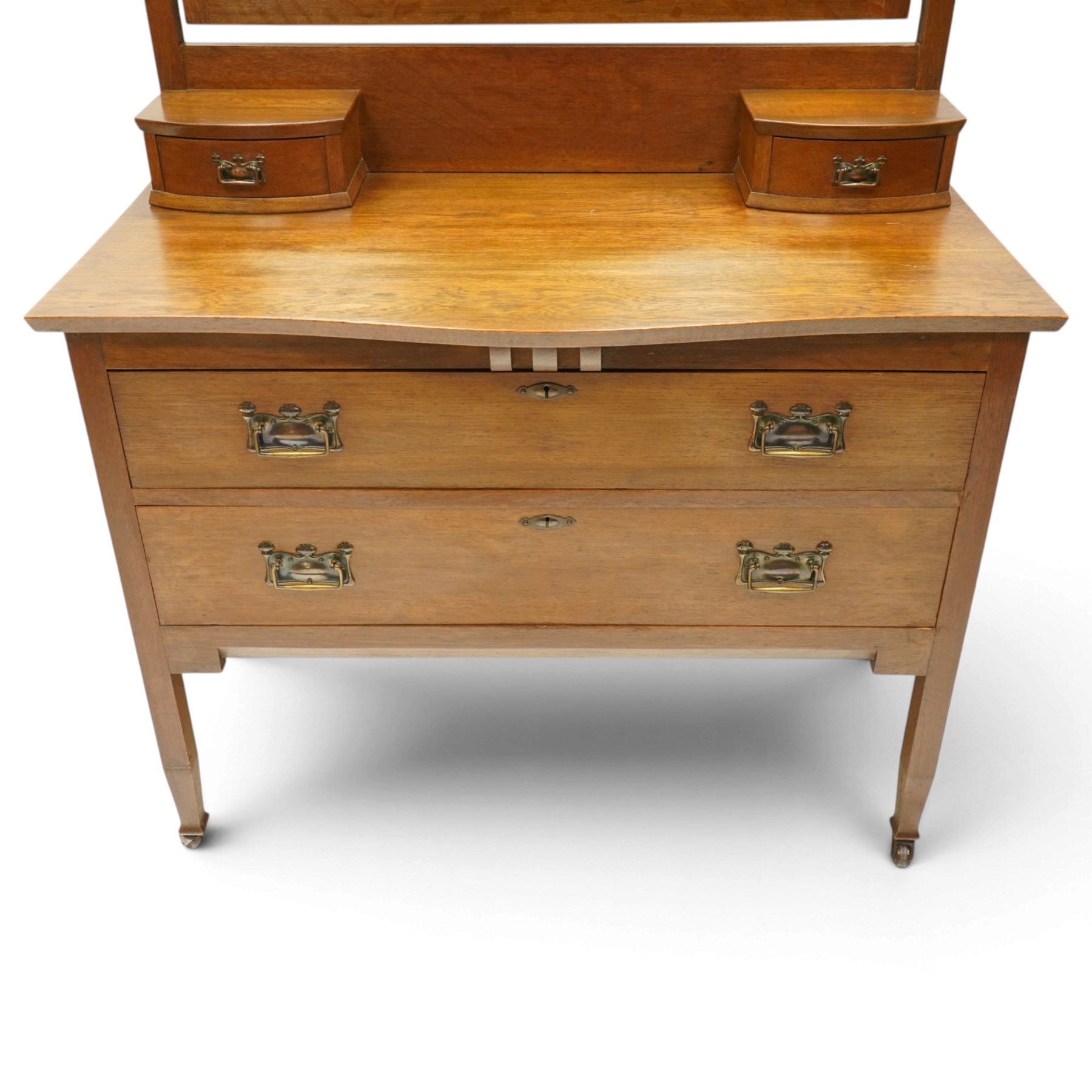 Manner of Liberty & Co. - Arts & Crafts oak dressing chest, the arched swing mirror with roundel-carved frieze and bevelled plate, serpentine-front top with two raised trinket drawers, fitted with two drawers with brass Art-Nouveau poppy handle-plates, on square tapering supports with castors