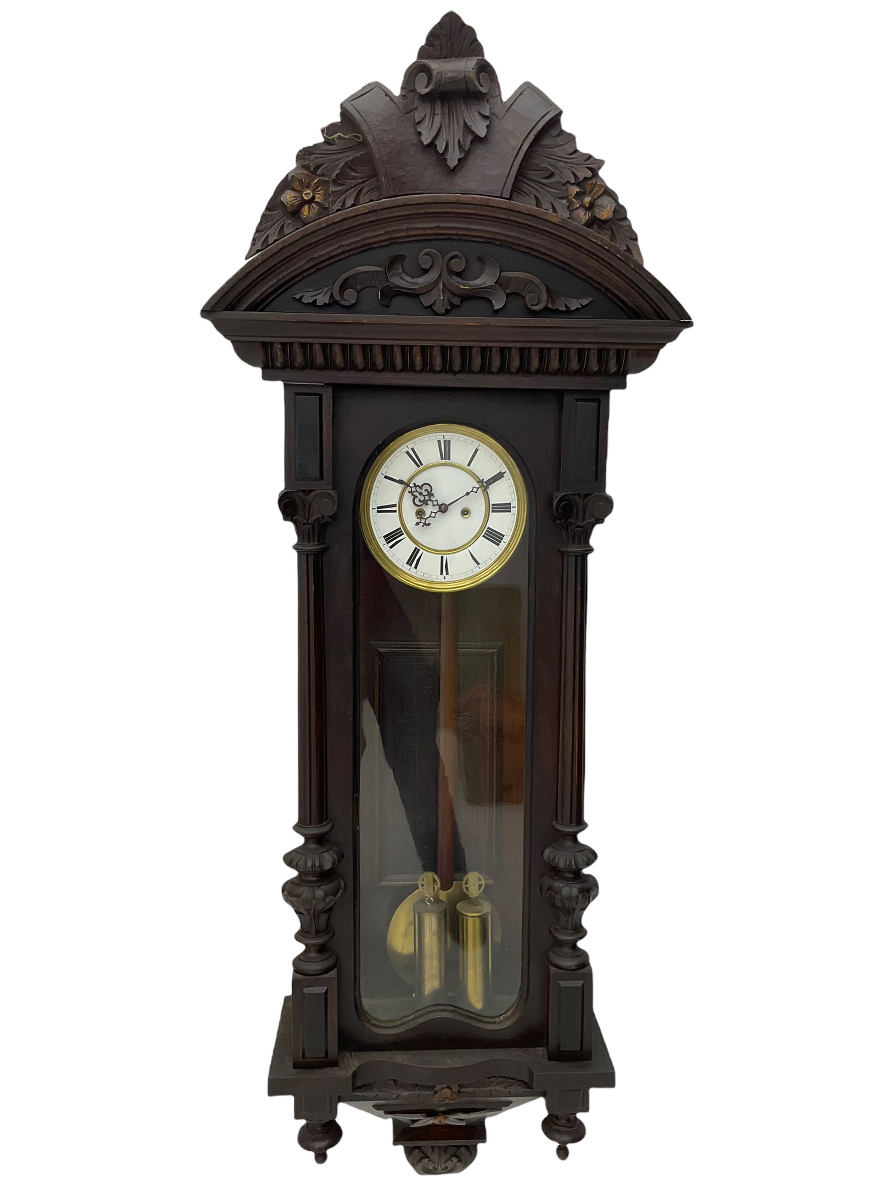 A late 19th century German Vienna style weight driven wall clock c1880, with a predominant arched pediment and applied carving, case with glass side panels (one missing) and a glazed full-length arched door flanked by reeded columns with decorative capitals, 7' two-piece white enamel dial with roman numerals, minute markers and pierced steel gothic hands, pendulum with a wooden rod, pressed brass bob and beat plate, eight-day weight driven movement striking the hours and half hours on a coiled gong. With brass cased weights.



