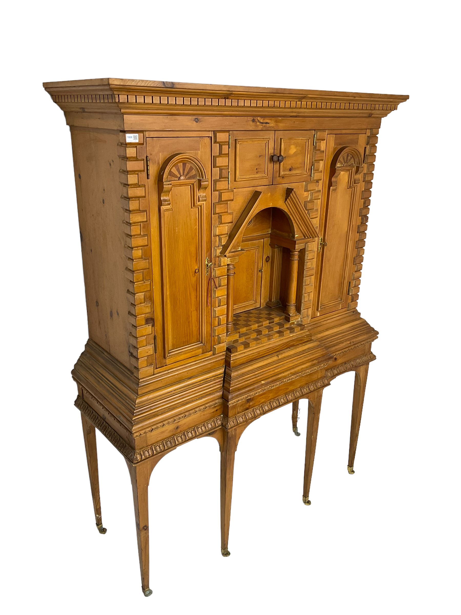 20th century pine architectural collector's cabinet on stand, projecting dentil cornice over various compartments, central recessed arch with turned pillar supports and sloped arch pediment, fitted with four cupboards, false brickwork uprights to front and corners, central hidden drawer revealing cube work surface, lower moulded plinth, the stand with foliate carved mouldings, on six square tapering supports with brass castors