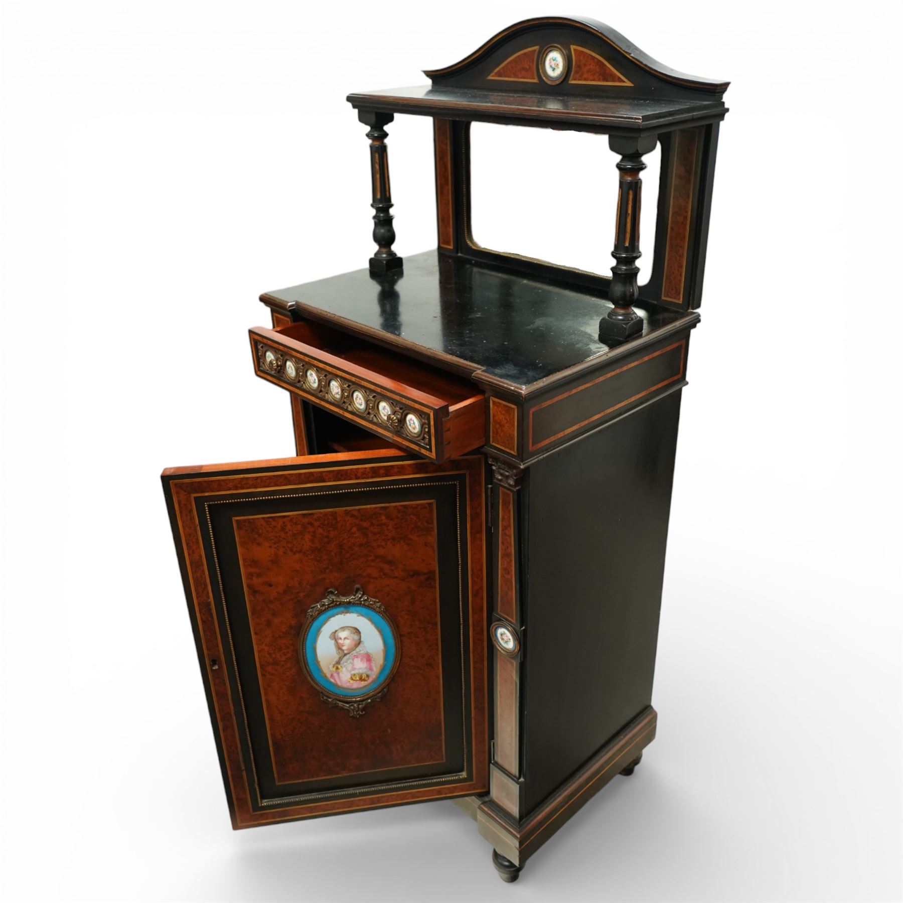 Victorian Aesthetic Movement amboyna and ebonised cabinet, the arched raised back inlaid with amboyna and floral porcelain plaque, over a raised shelf with fluted and turned supports, and a bevelled mirror plate with brass beading beneath, the inverted-breakfront top over an ornate frieze drawer with guilloche style gilt-metal mounts and porcelain plaques, the panelled door with a central porcelain plaque depicting a lady in a fuchsia gown within concentric ebonised and amboyna inlays, flanked by architectural style uprights with Corinthian capitals, raised on turned feet