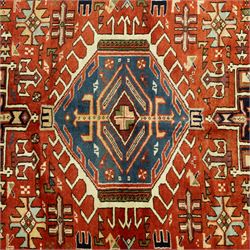 North West Persian Karajeh hand-knotted rug, terracotta ground, with a stepped geometric pole medallion design in blue, ivory and dark navy tones, surrounded by angular tribal motifs incorporating stylised flowerheads, hooked devices and geometric vine work, enclosed by a dark navy border with repeating rosettes and angular leaf motifs, flanked by narrow patterned guard stripes, with cotton fringes