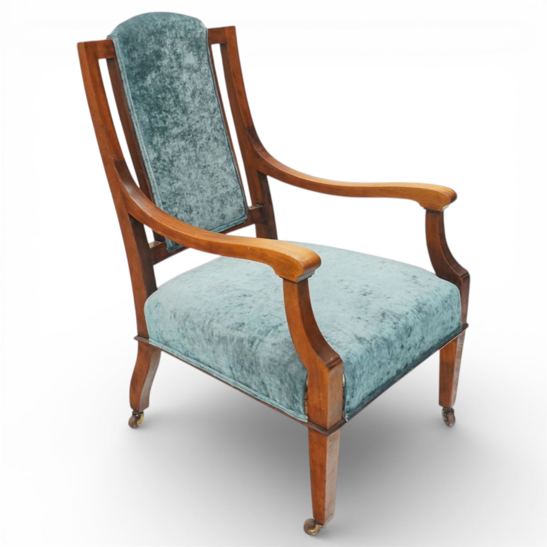 Arts & Crafts mahogany framed armchair, shaped rectangular back with open uprights, padded back, seat and arm supports upholstered in blue velvet, downswept arms on square tapering front supports with brass caps and castors, the rear supports slightly splayed