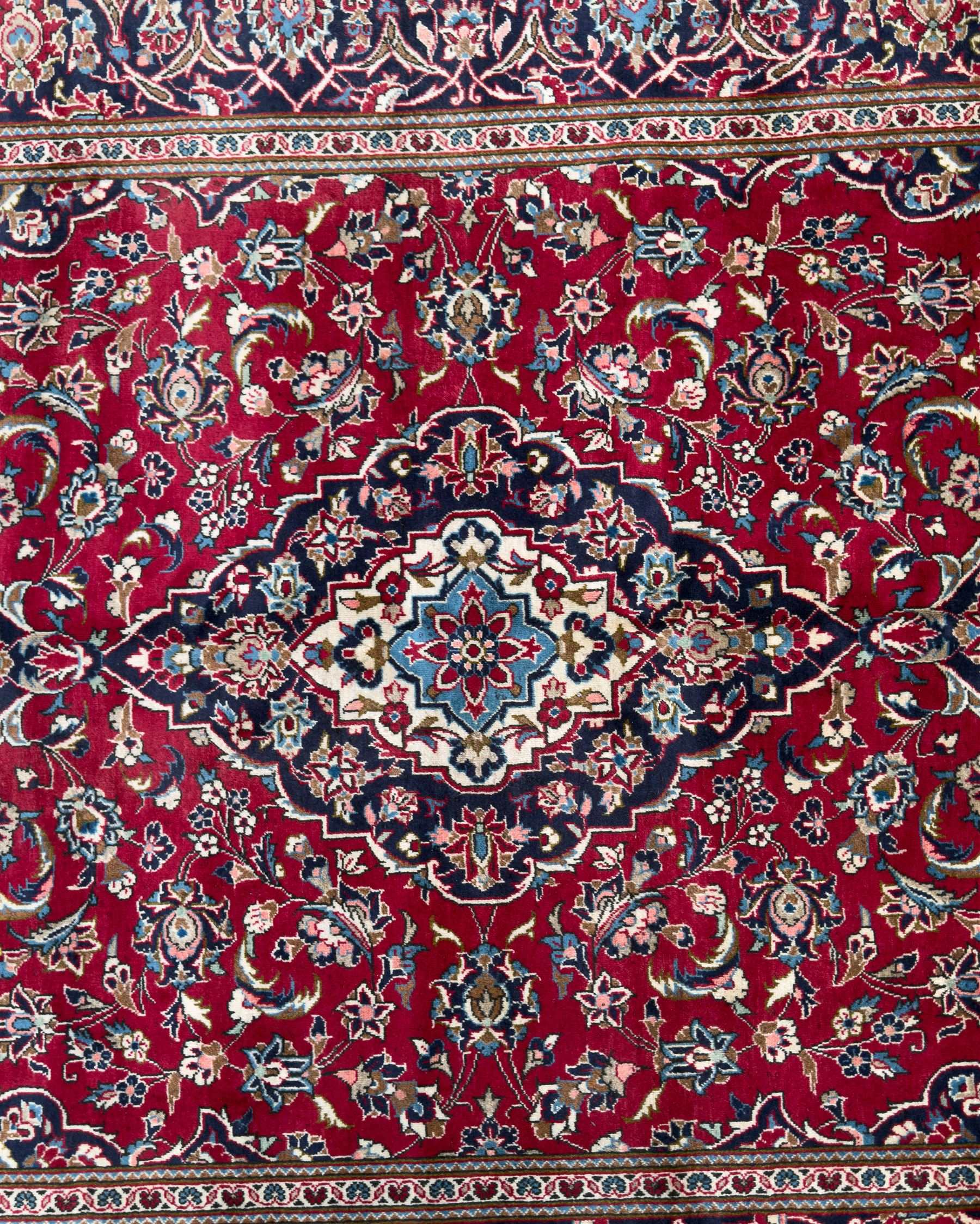 Central Persian Kashan hand-knotted carpet, the deep red ground with a central lozenge medallion in ivory and navy, surrounded by dense scrolling floral and vine motifs, navy spandrels and a wide border with stylised palmettes and flowerheads enclosed by multiple guard stripes
