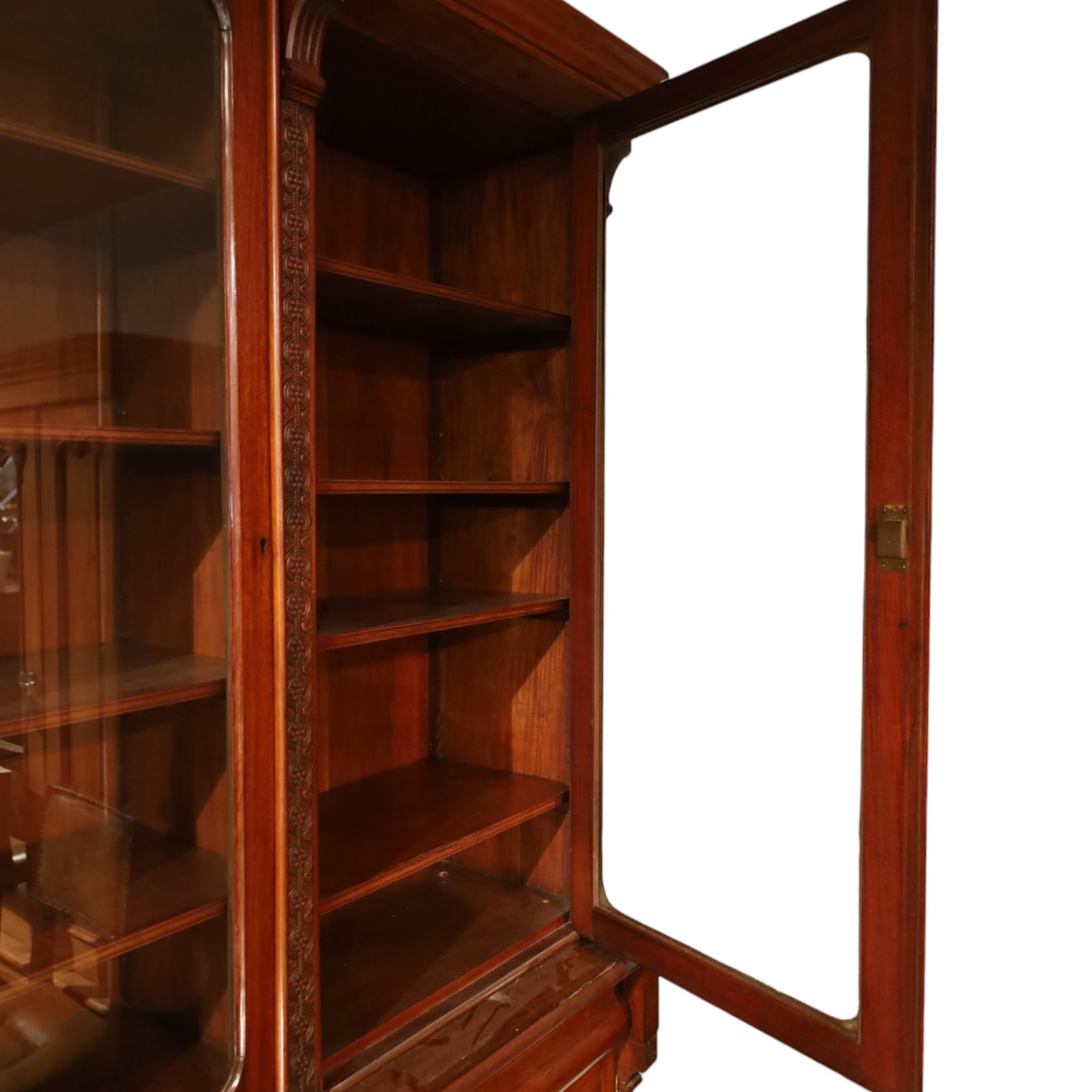 Victorian carved mahogany enclosed bookcase on cupboard, projecting moulded cornice over twin glazed doors enclosing adjustable shelves, flanked by foliate carved uprights, the lower section fitted with two cushion moulded drawers over twin panelled cupboard doors, divided and flanked by conforming carved uprights, on plinth base