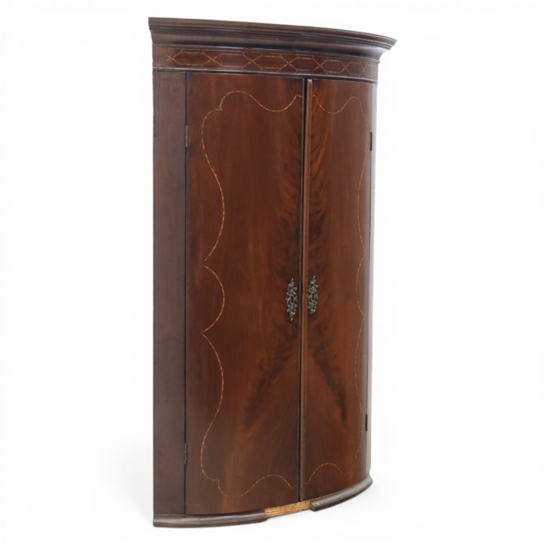 George III mahogany bow-front corner cupboard, projecting moulded cornice over satinwood inlaid frieze with geometric crossbanding, enclosed by twin shaped panel doors with line inlay and ornate brass escutcheon plates, painted interior fitted with shaped shelves, on moulded plinth base