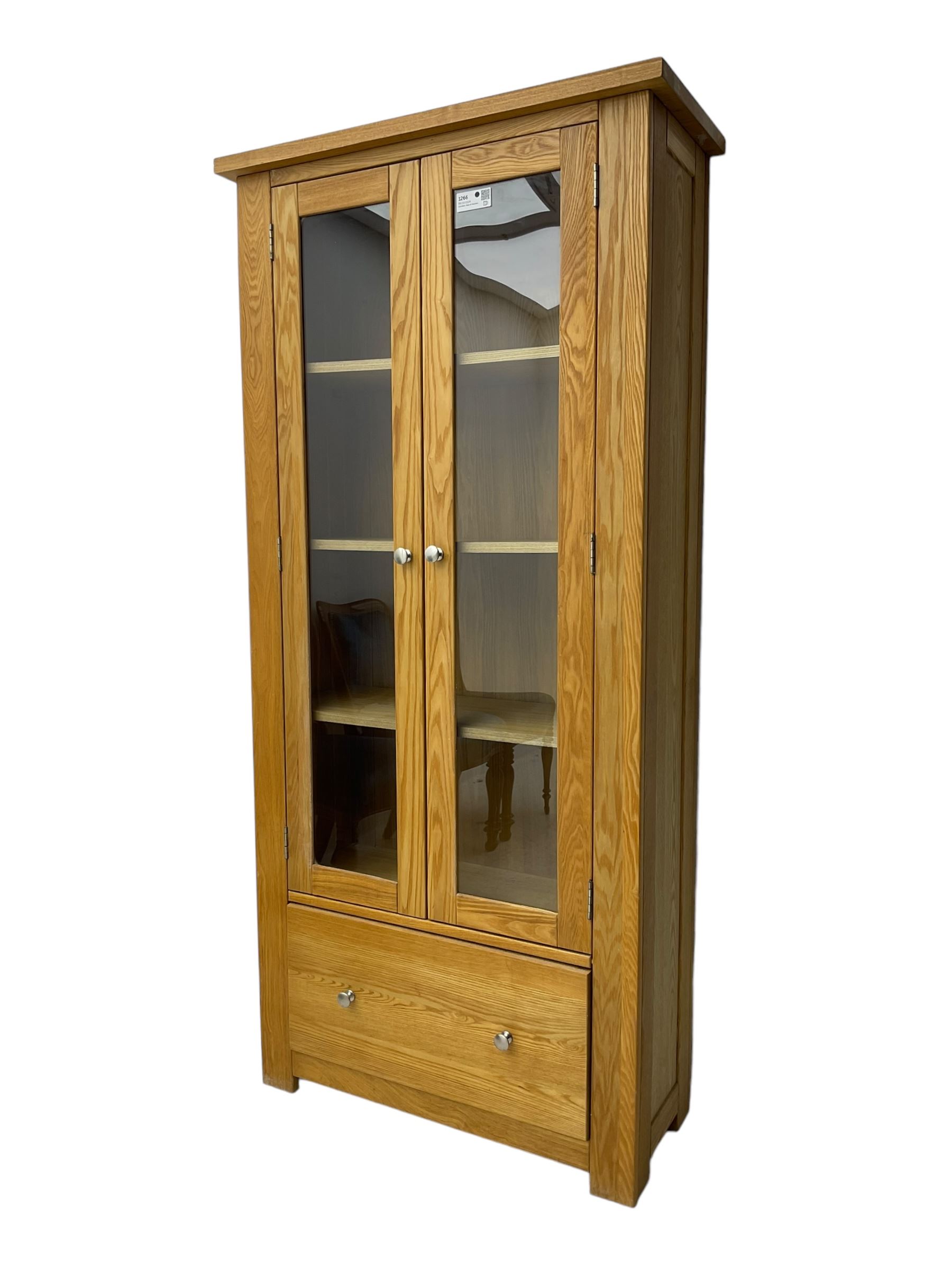Light oak bookcase display cabinet, projecting square cornice over glazed double doors enclosing three adjustable shelves, single drawer base, on stile supports