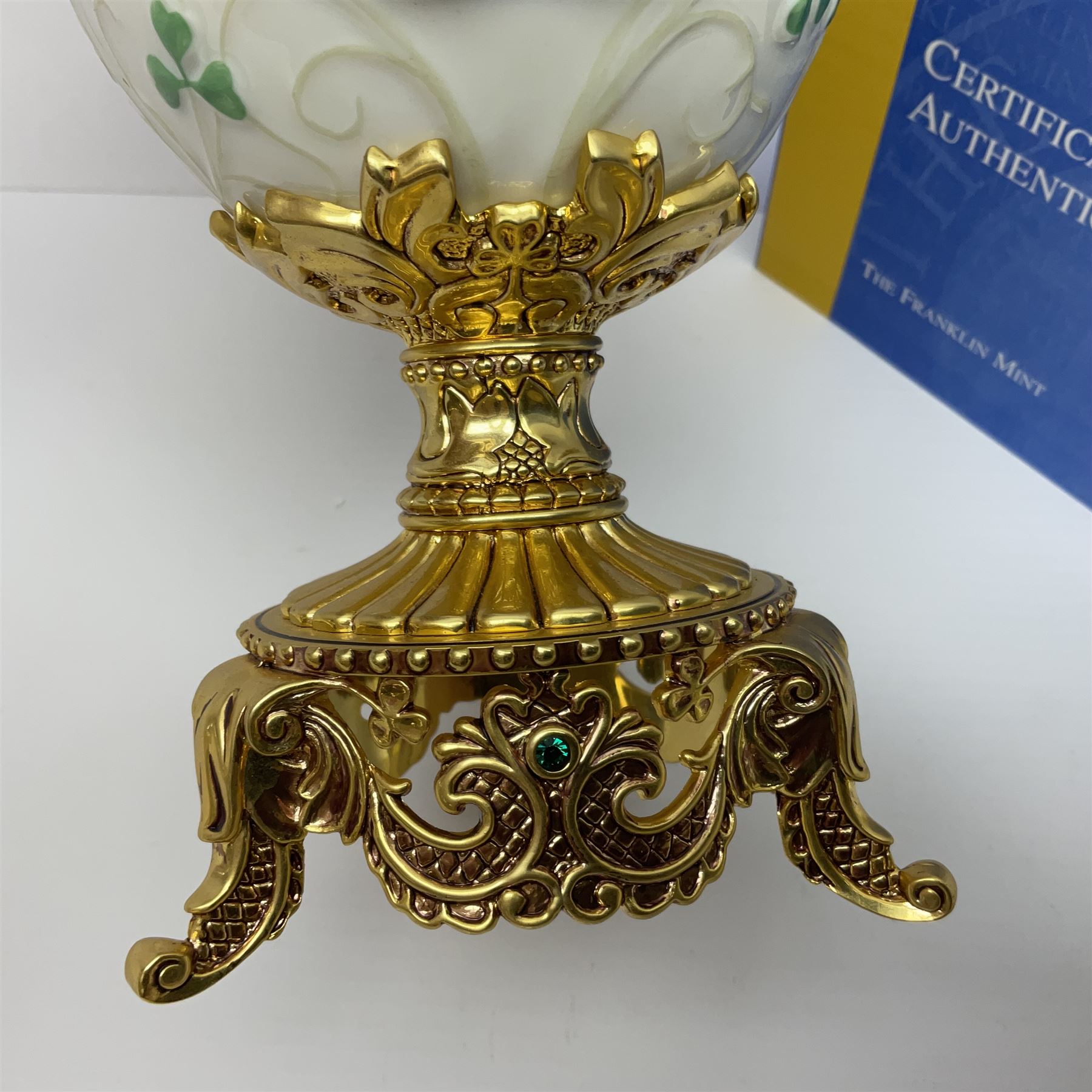 Franklin Mint The Emerald Isle Collector Egg, the egg decorated with embossed shamrocks upon a cream ground, with a gilded shamrock finial, accents and openwork door, opening to reveal a miniature landscape scene, upon three gilded feet set with green paste stones, with certificate of authenticity H23cm, 