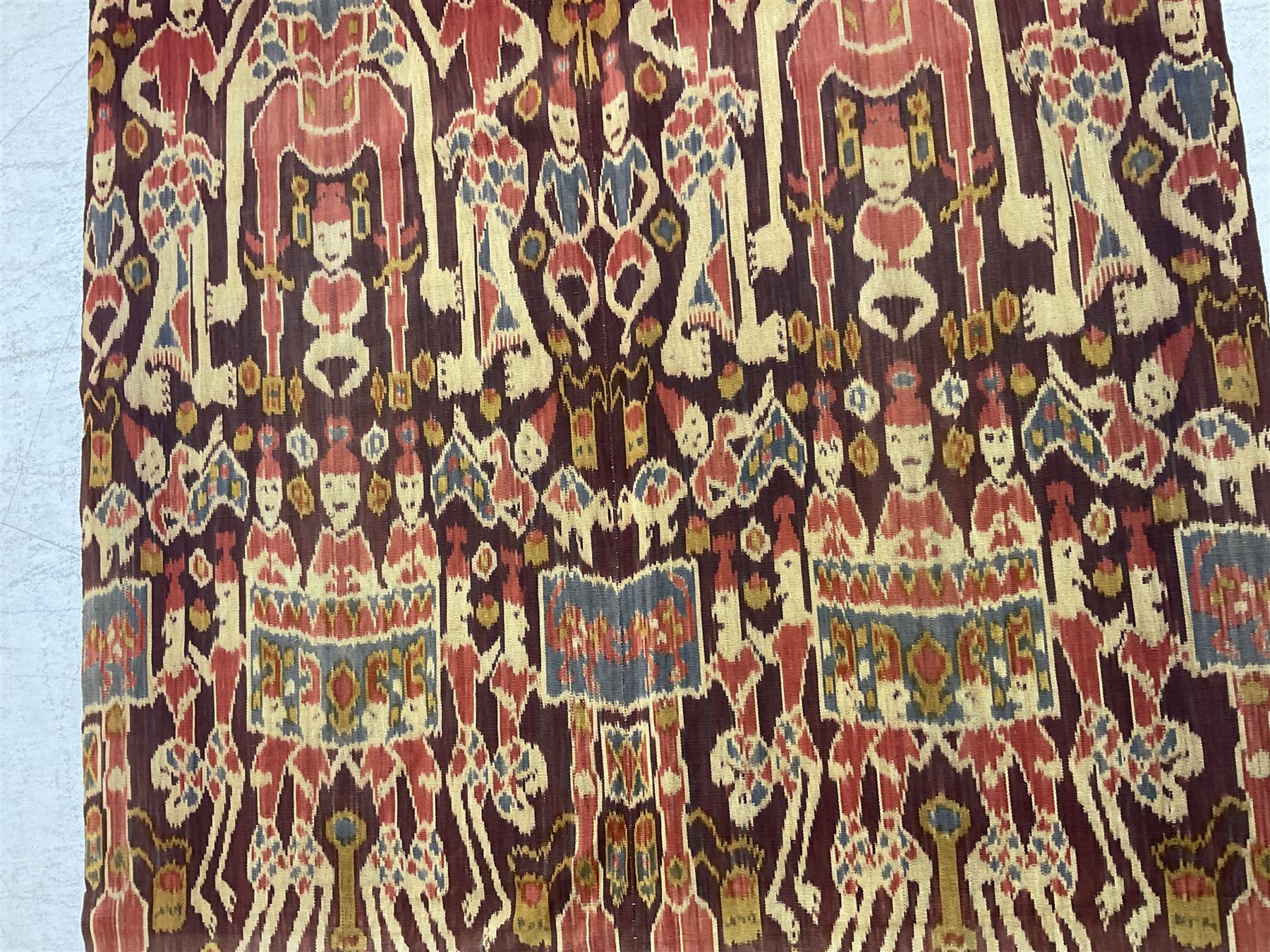 Ikat Man’s Cloth (Hinggi Kombu), East Sumba, Indonesia, woven in red, blue and yellow-brown warp ikat, constructed of two mirroring long panels, the motifs arranged in four bands depicting warrior figures astride rearing horses, high-ranking figures and guardians within geometric borders, on carved wooden wall hanger, L295cm
