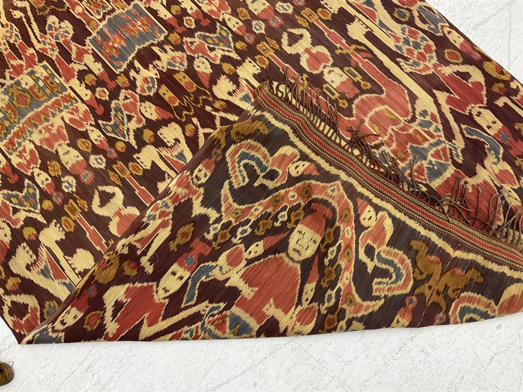 Ikat Man’s Cloth (Hinggi Kombu), East Sumba, Indonesia, woven in red, blue and yellow-brown warp ikat, constructed of two mirroring long panels, the motifs arranged in four bands depicting warrior figures astride rearing horses, high-ranking figures and guardians within geometric borders, on carved wooden wall hanger, L295cm
