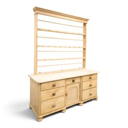 Victorian stripped pine dresser base with plate rack, the four-tier plate rack with supporting rails and hooks, the inverted-breakfront base fitted with seven assorted graduating drawers surrounding a recessed cupboard, on compressed bun feet with castors
