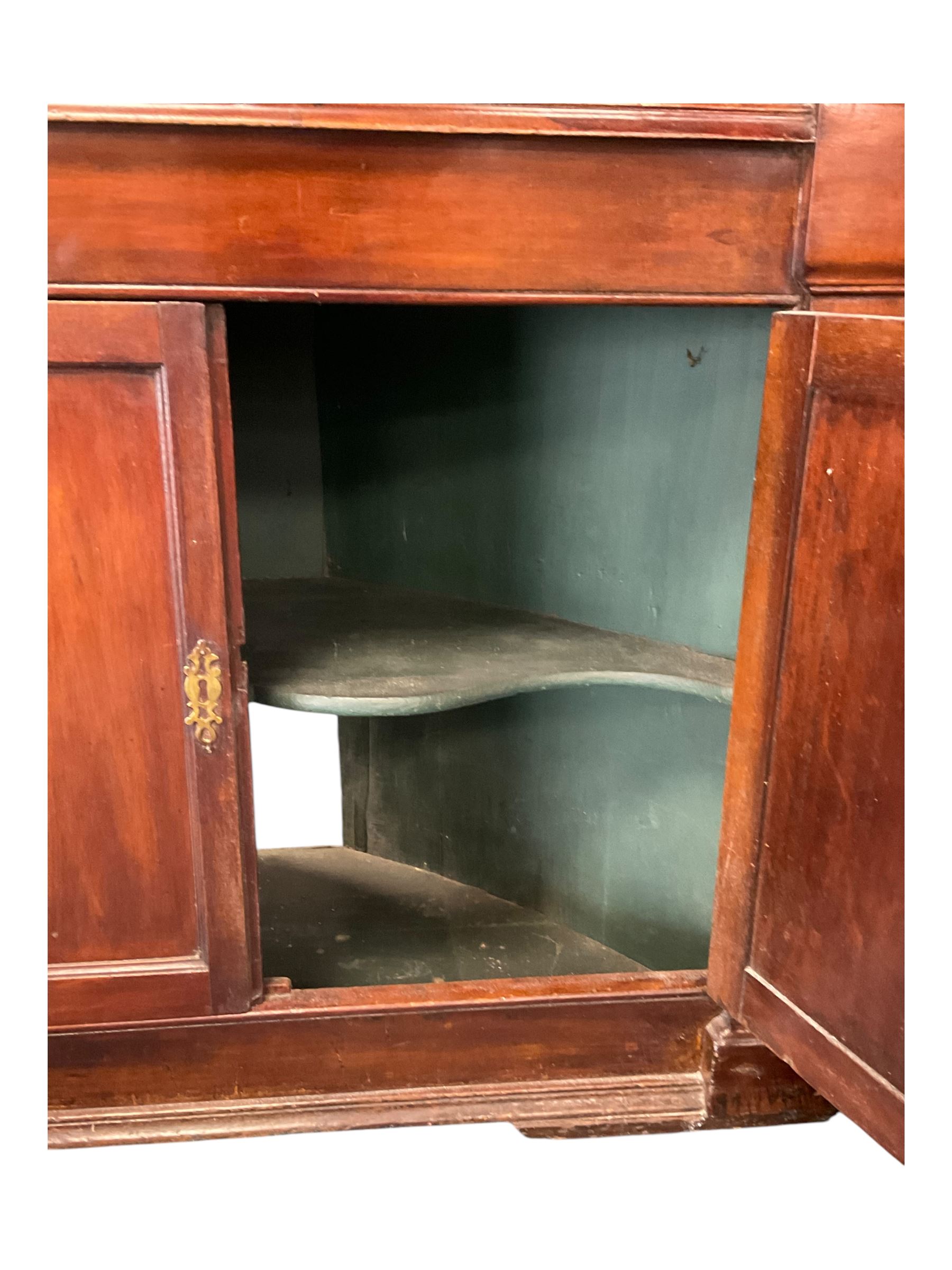 George III mahogany corner cabinet, the scrolled pediment surmounted by a brass finial and carved fretwork frieze, with fluted uprights flanking arched double doors over a painted interior fitted with shaped shelves, above a lower cupboard with twin panelled doors, on plinth base