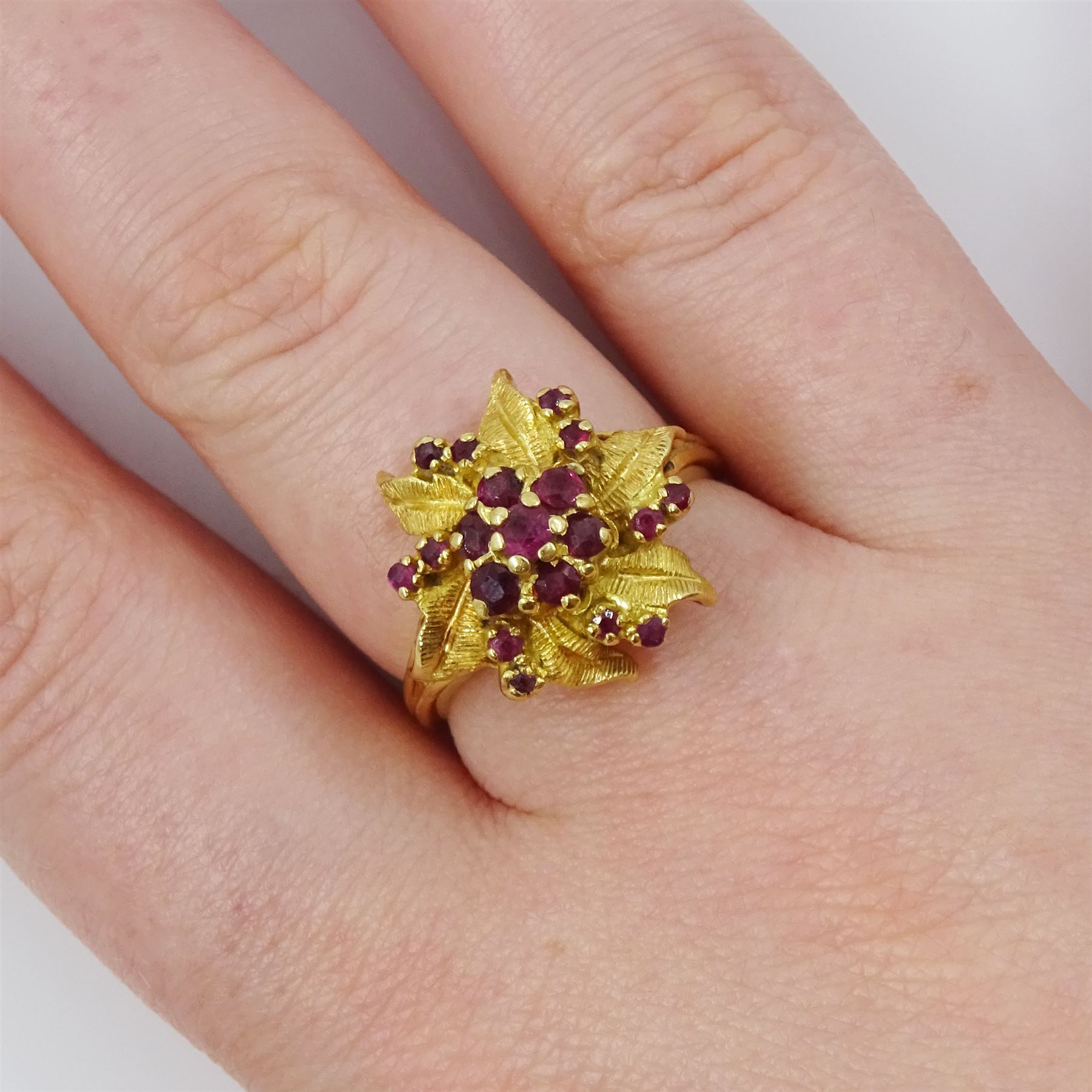 18ct gold ruby flower head cluster ring, seven round cut rubies surrounded by textured leaves, each spaced by two rubies, stamped 750