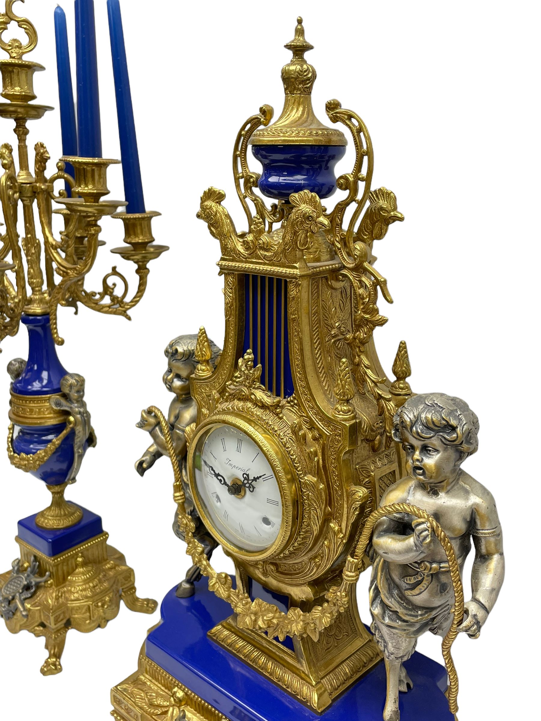 A 20th century continental gilt metal Lyre mantle clock with a pair of matching six light candelabra, spring driven movement housed in a gilt drum case on a brass and cobalt blue base with paw feet, two bronzed metal figures of mythological cherubs with animal legs and cloven hooves supporting a festooned garland swag, eight-day twin barrel striking movement with a floating lever balance escapement, striking the hours and half-hours on two bells, white enamel dial with roman numerals, minute track and pierced steel hands, dial inscribed “Imperial”. 