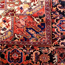 North West Persian Heriz hand-knotted carpet, madder red field with large central stepped medallion with angular floral and geometric designs, surrounded by stylized palmettes, leaves and tribal motifs with indigo corner spandrels, enclosed by a broad navy border of geometric floral devices between multiple guard stripes, with cotton fringe ends 
