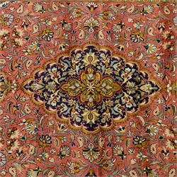 Central Persian Qom hand-knotted carpet, salmon pink ground, with a central dark navy shaped floral medallion with pendants, surrounded by dense scrolling foliate and floral motifs incorporating rosettes, palmettes and delicate vine work, enclosed by ivory spandrels and a dark navy border with repeating palmettes and meandering vines, flanked by narrow floral guard stripes, with cotton fringes