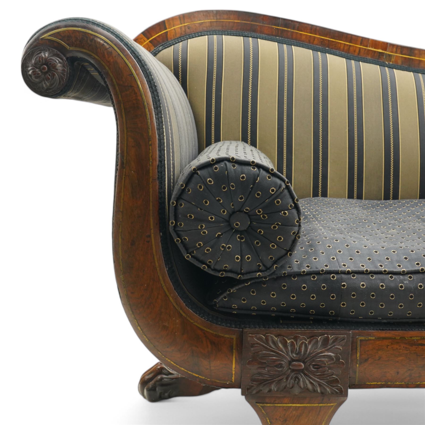 Regency brass inlaid rosewood chaise lounge or settee, shaped back over rolled head and foot, upholstered in black and gold striped fabric with rectangular squab cushion and bolster cushion, the rolled terminals carved with acanthus leaf roundels, lower rectangular foliate carved panels, on out splayed supports with paw carved feet 