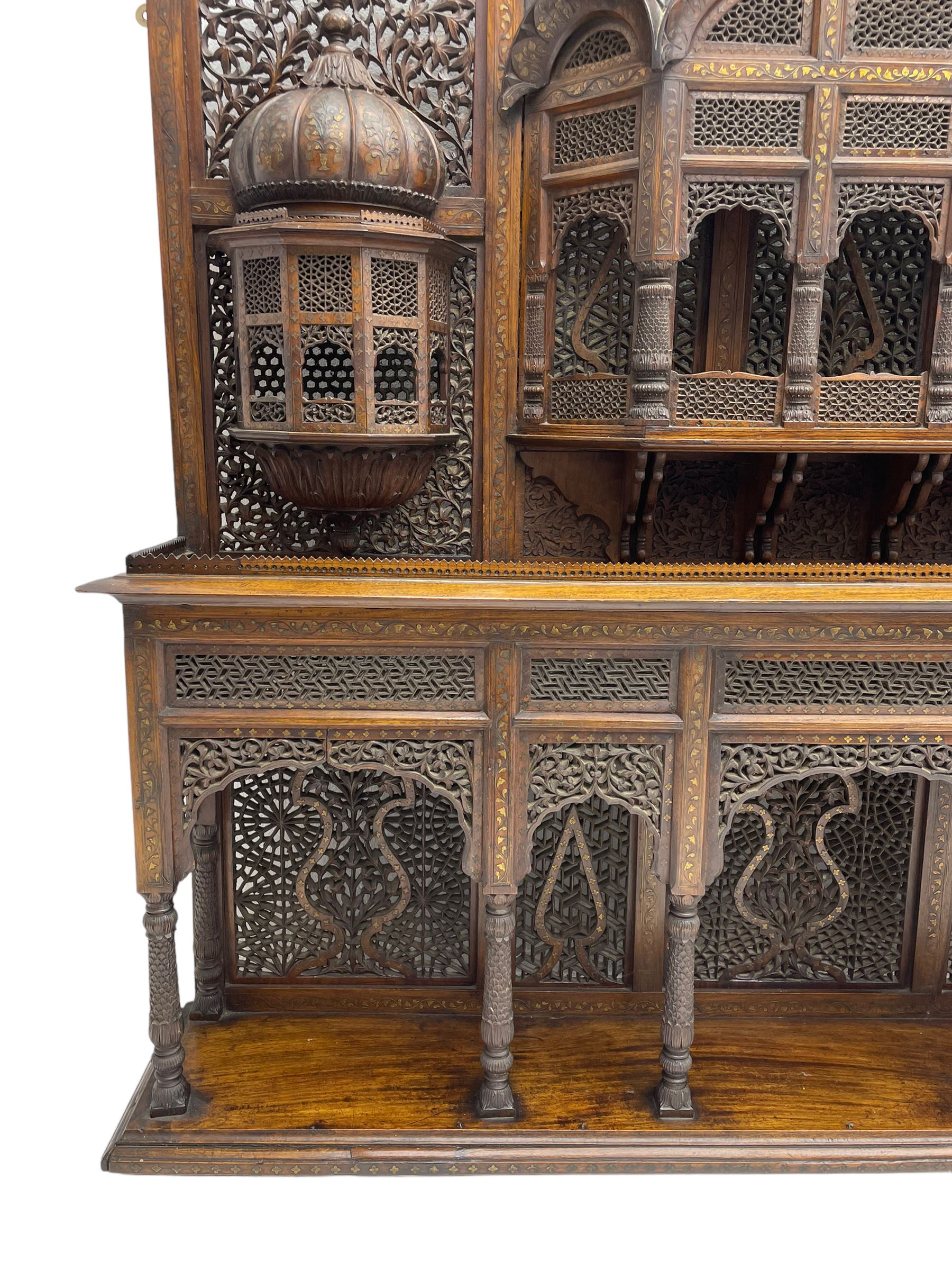 Late 19th century Indian Burmese carved and pierced hardwood and brass inlaid wall shelf, the shaped back profusely decorated with trailing and interlaced foliage, with projecting architectural structures, the central balcony with canopy top and five open windows with arched apertures on columns, two flanking five-sided balconies, the shelf supported by six foliate carved columns with pierced cusped pointed arches 