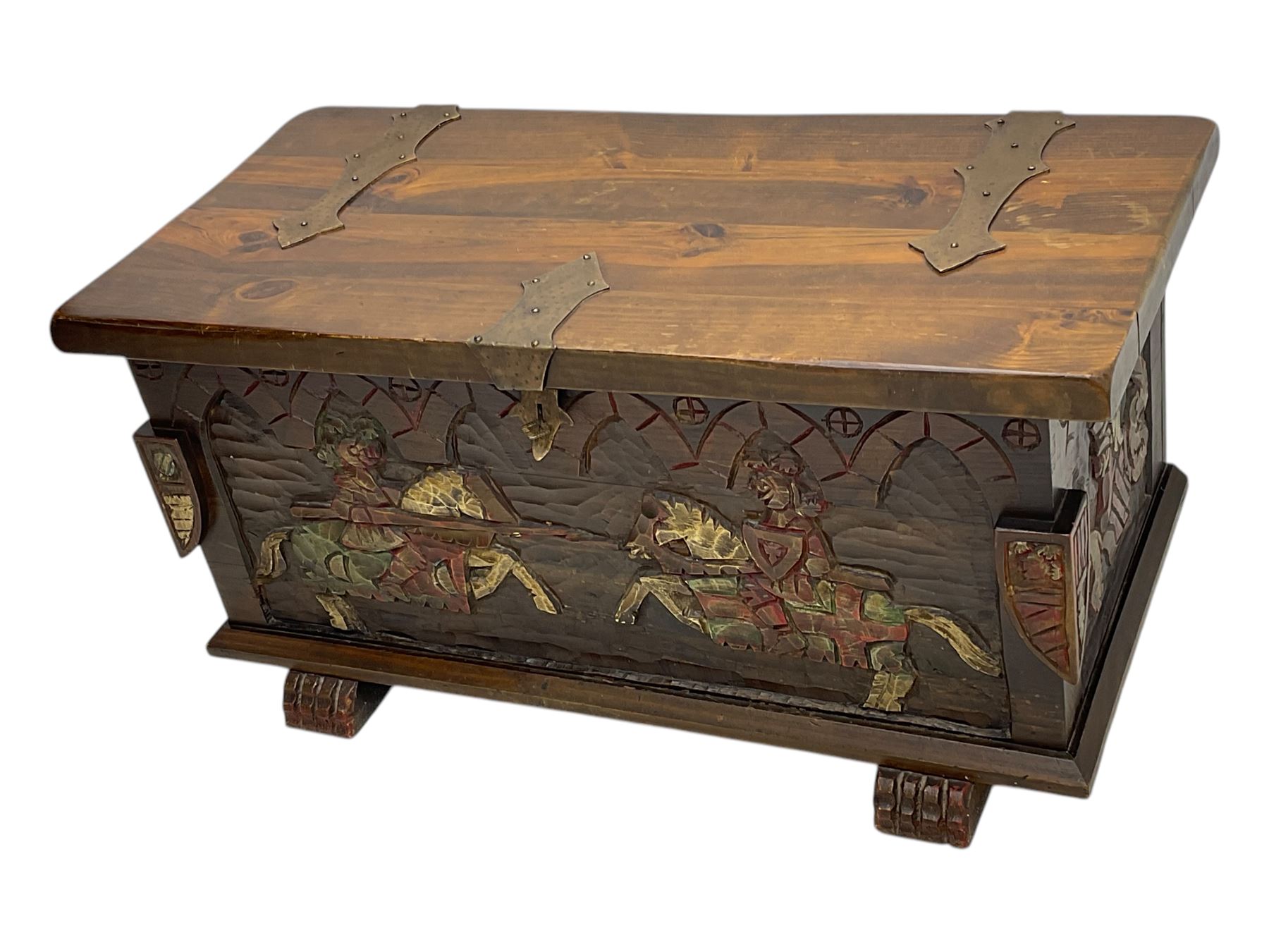Mid-20th century carved hardwood chest, the rectangular hinged top with decorative strap hinges, the front and sides deeply carved with medieval-style jousting knights, heraldic shields, and architectural elements, raised on shaped block feet