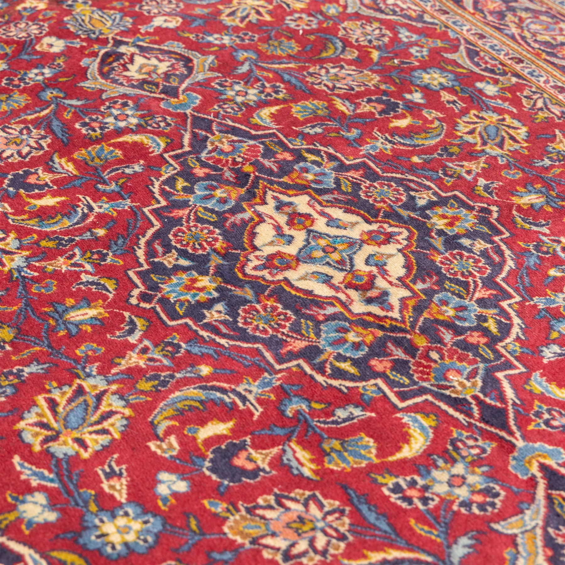 Central Persian Kashan hand-knotted rug, crimson field, profusely decorated with scrolling vines and palmettes, floral sprays around an ivory and indigo lobbed medallion enclosed by spandrels within an indigo border of arabesques between multiple guard stripes  