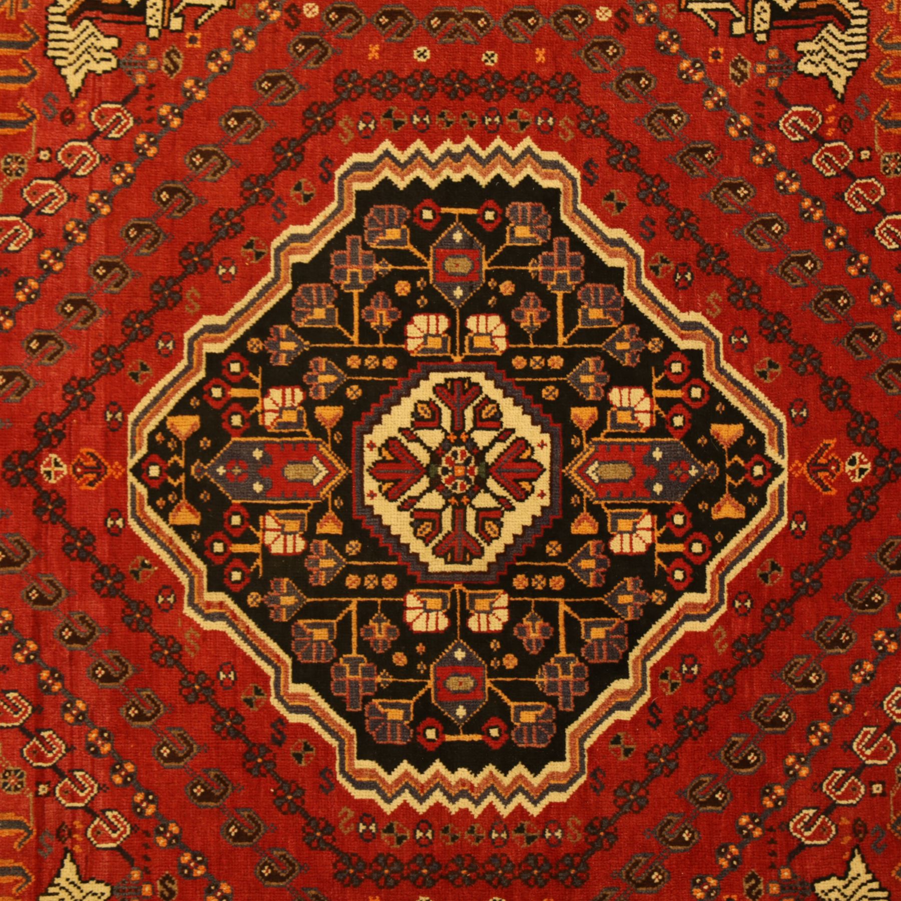 Persian Shiraz rug, hand-knotted tribal rug, bold geometric central medallion, set within a large hexagonal crimson field with angular stepped corners in dark blue, with boteh motifs, rosettes, and other tribal symbols arranged in diagonal rows, the corners with indigo spandrels decorated with zoomorphic and arboreal motifs, main border composed of a floral meander motif with large rosette, flanked guard borders with vine and leaf motifs 