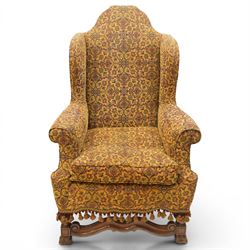 Pair of William & Mary design wingback armchairs, the arched high-back over rolled arms with a loose seat cushion and sprung seat, upholstered in old-gold ground foliate patterned fabric, raise on scroll feet united by a front serpentine stretcher
