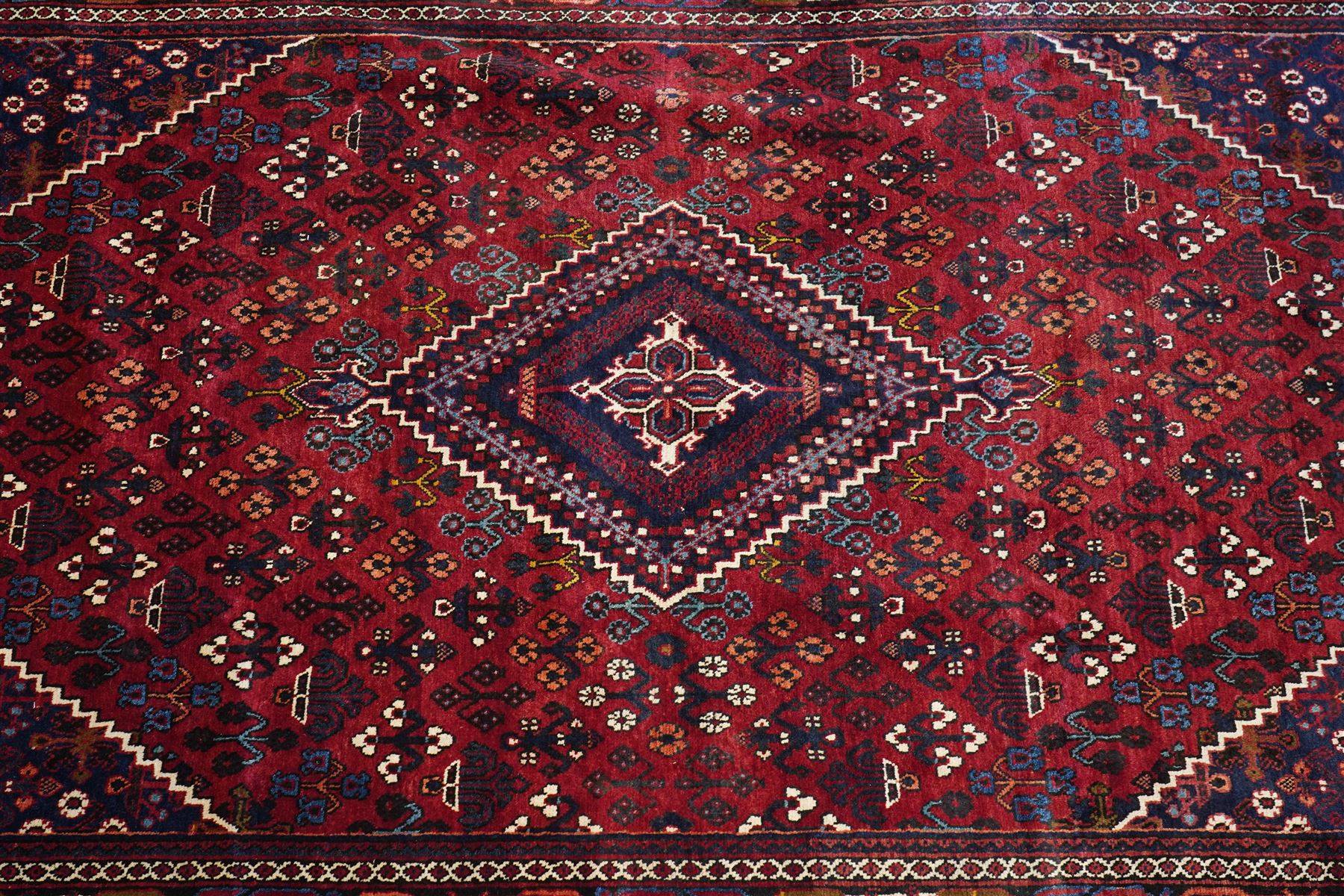 Central Persian Joshaghan hand-knotted carpet, deep red field decorated with a lattice design of stylized boteh and palmette motifs arranged around a stepped diamond medallion, navy corner spandrels similarly decorated enclosed by a dark indigo main border of floral and geometric motifs between multiple guard stripes, with cotton fringe ends 