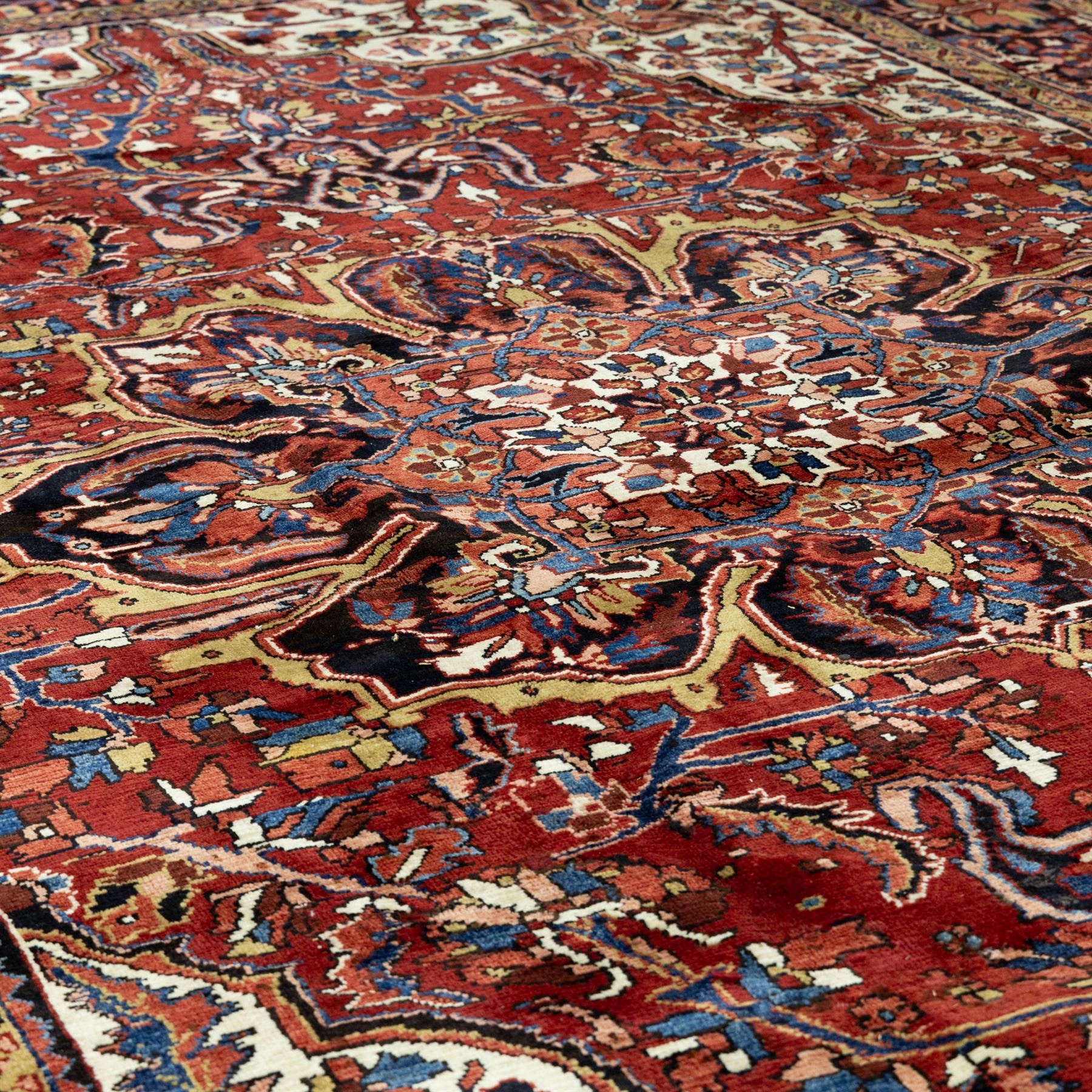 North West Persian Ahar hand-knotted carpet, madder red ground, with a large central shaped floral medallion with pendants, surrounded by angular scrolling foliate motifs incorporating rosettes, palmettes and stylised flowerheads, enclosed by ivory spandrels and a dark navy border with repeating geometric floral motifs and meandering vines, flanked by narrow patterned guard stripes, with cotton fringes