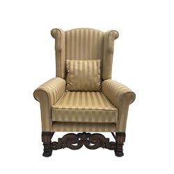 William and Mary style wingback armchair, upholstered in gold striped fabric, on walnut scroll carved supports joined by scroll and foliate carved middle rail, turned H-shaped stretchers, on castors