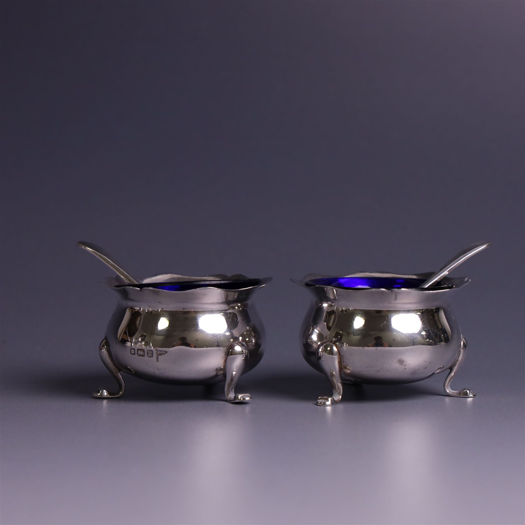 Early 20th Century four piece silver cruet set, comprising a pair of pepper shakers and a pair of open salts, each of circular form with shaped rims and upon three pad feet, hallmarked Walker & Hall, Sheffield 1912, the open salts with two silver plated salt spoons and blue glass liners 